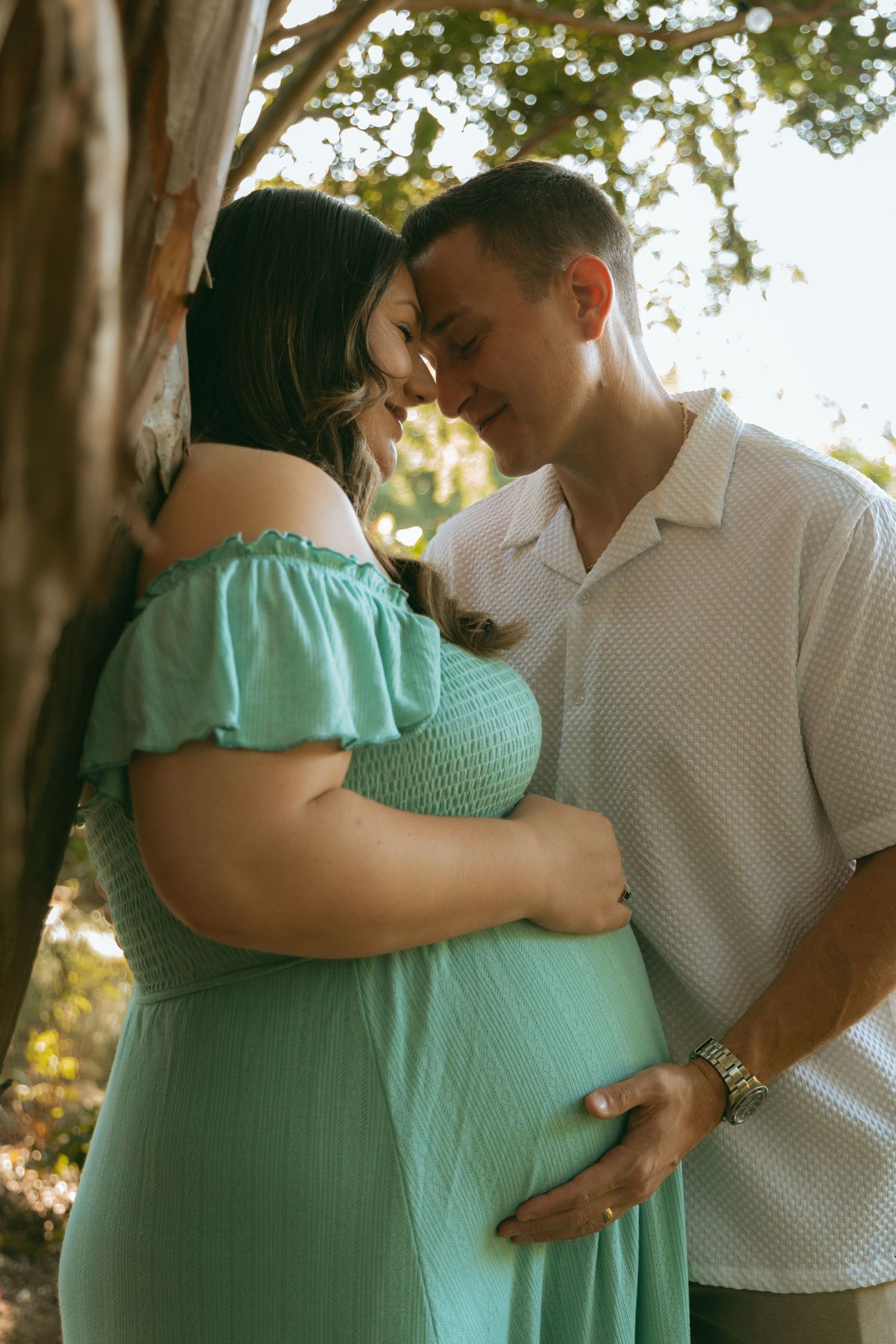 Couple sharing a tender moment outdoors, pregnant woman in teal off-shoulder dress, man in white shirt holding her belly.