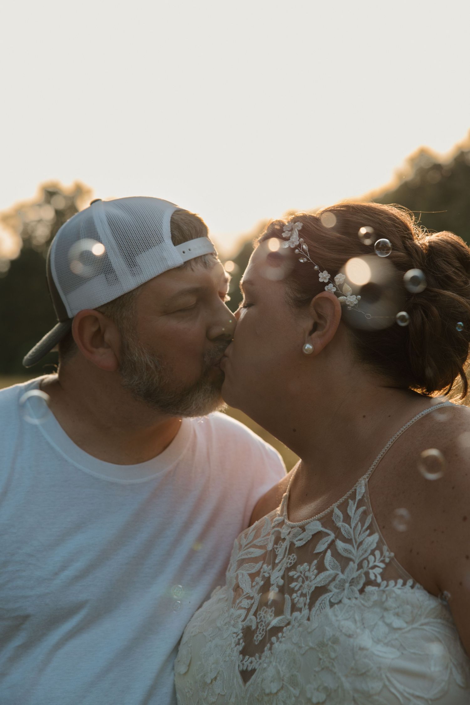 Couple sharing a romantic kiss at golden hour, bride in lace dress with floral hair piece, groom in white tee and cap.