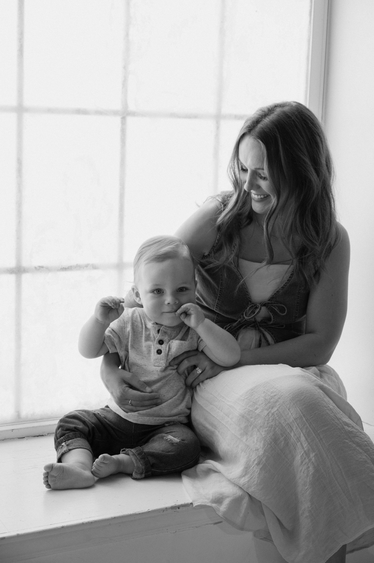 Black and white photo of a smiling mother sitting by a window with her happy toddler on her lap.