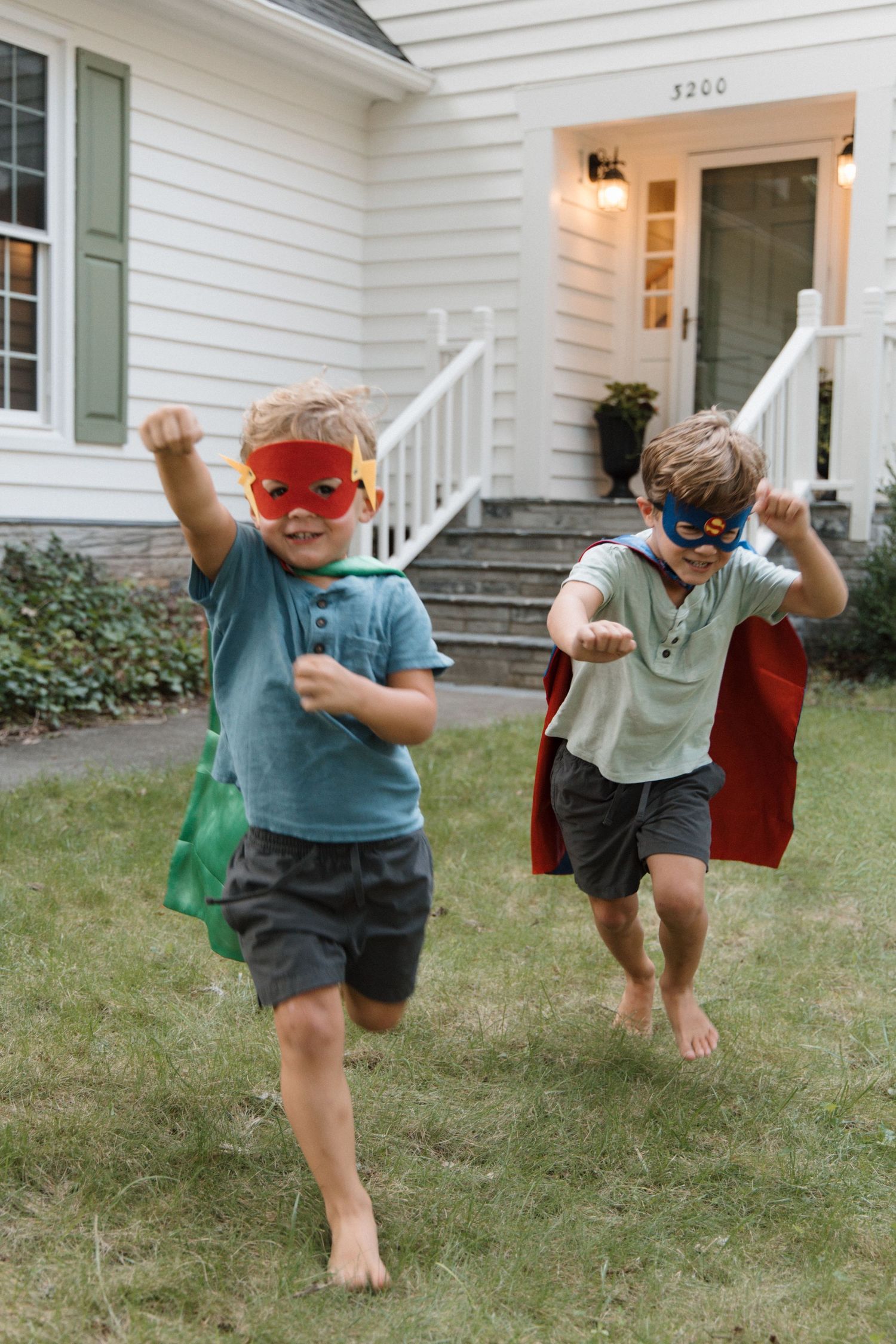 Two boys in superhero capes and masks run barefoot on grass in front of a white house.