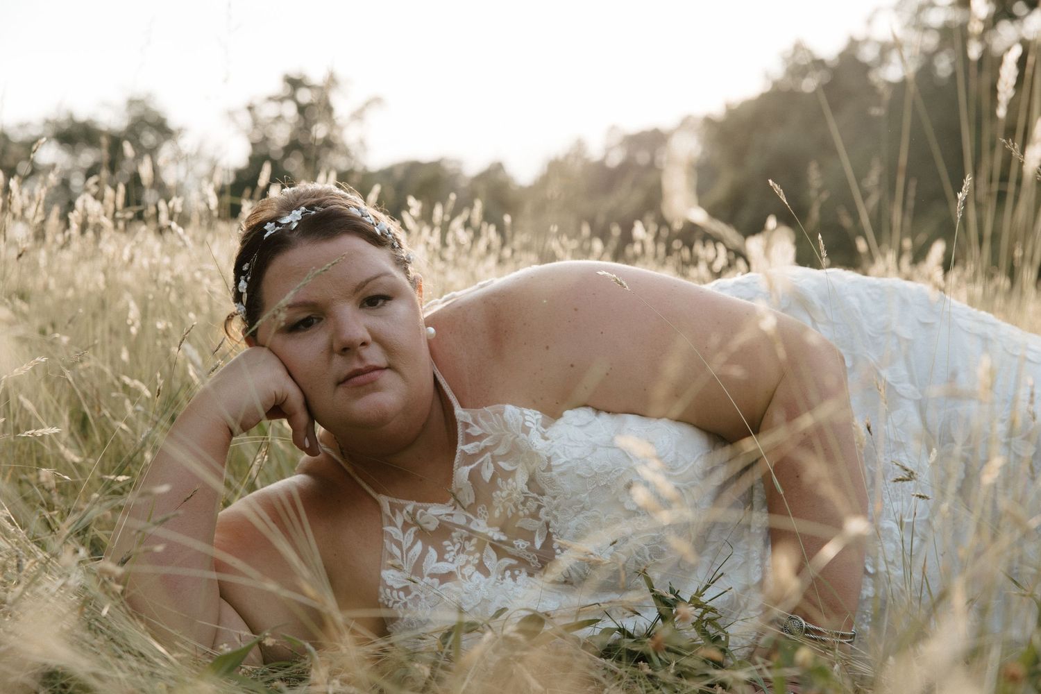 Bride in white gown lying in golden field grass, wearing hair accessories, in a dreamy outdoor wedding photo.