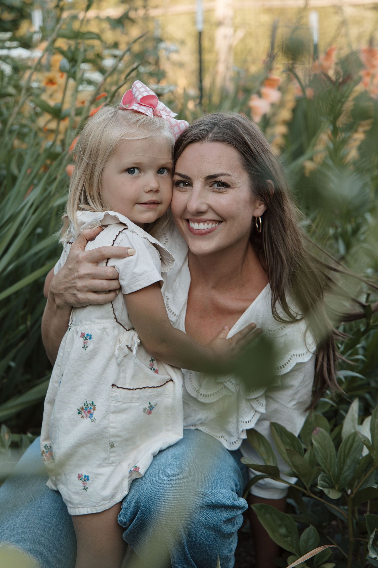 A smiling woman holds a young blonde girl with a pink bow in a lush garden setting.