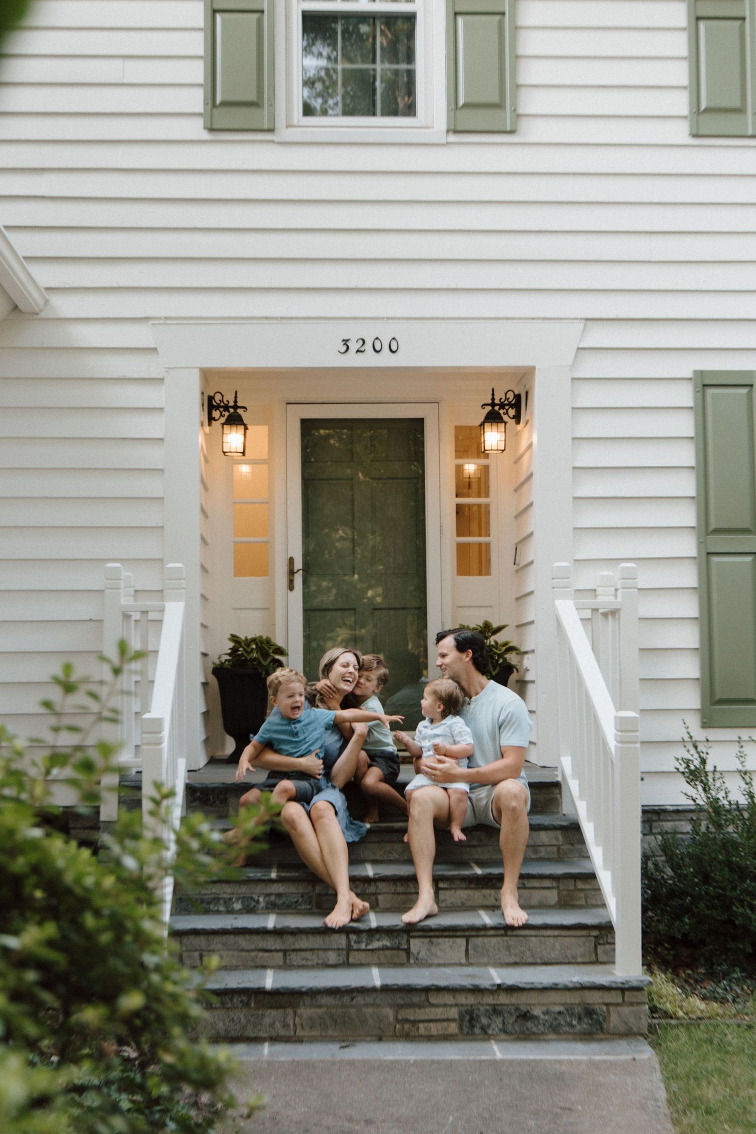 Family of four sitting barefoot on front porch steps of white home with green door, sharing a joyful moment together.