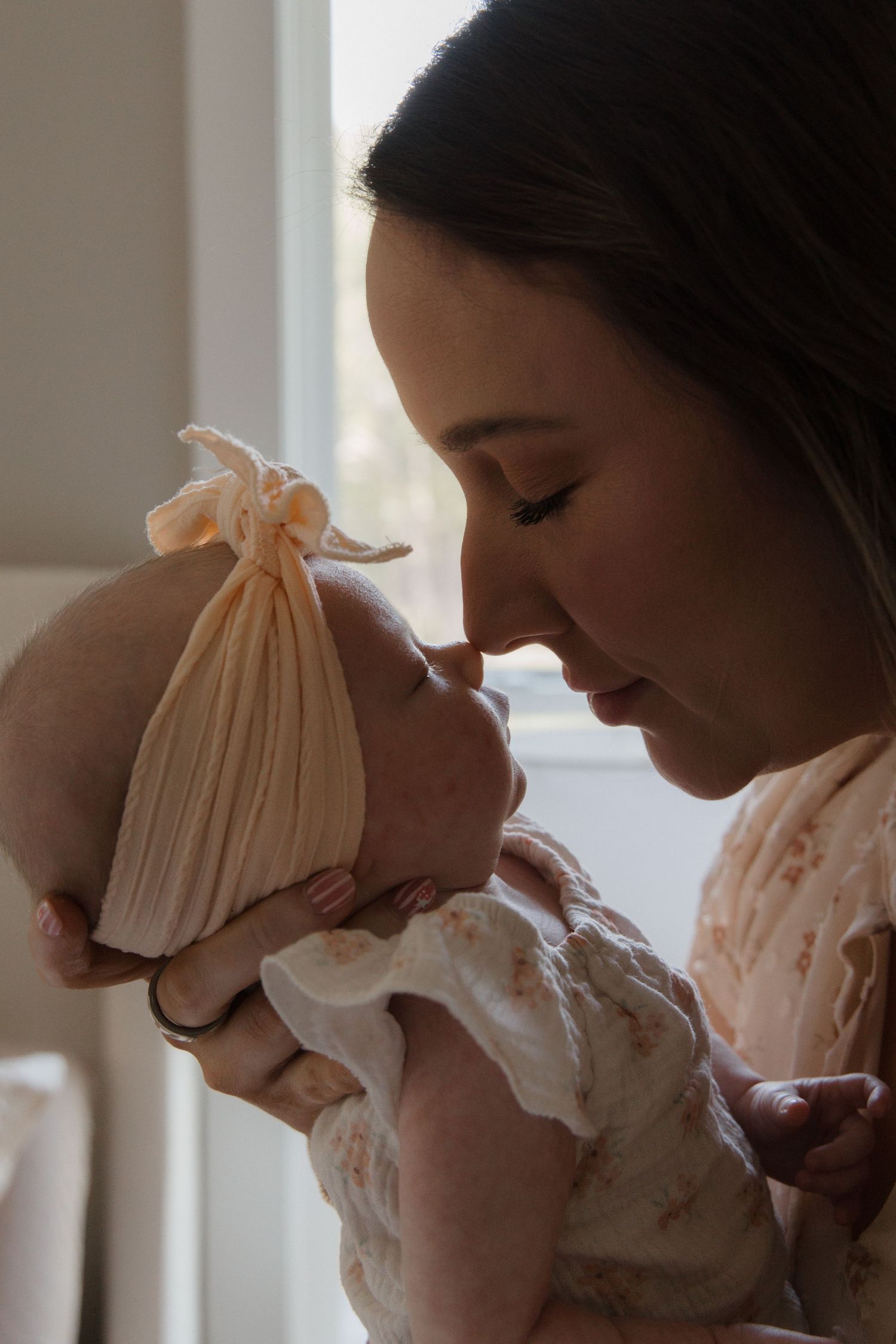 A mother tenderly touches noses with her newborn baby wearing a cream knot hat, sharing a sweet bonding moment.