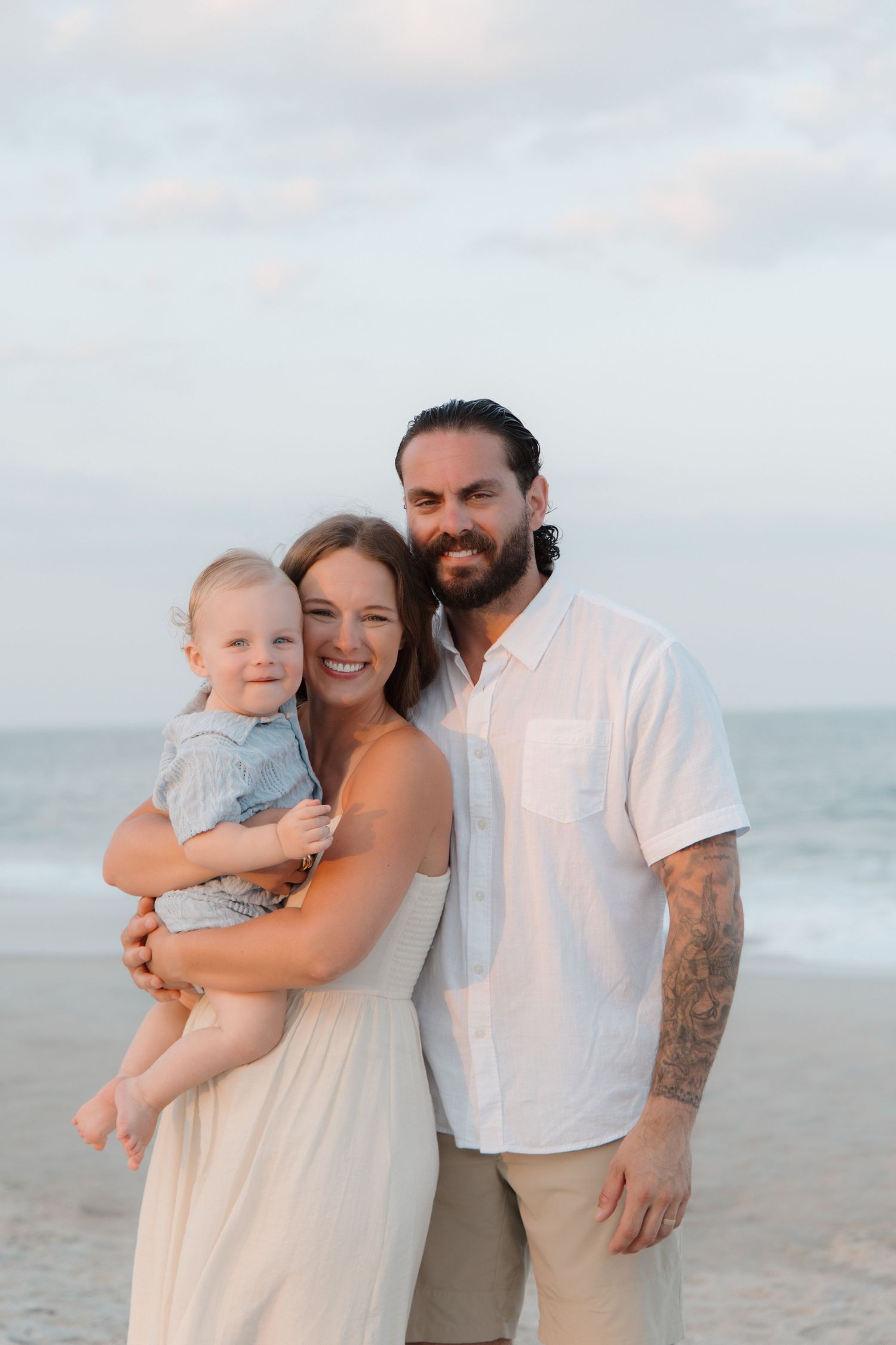 A smiling family of three poses on a beach at sunset, with mom holding a baby and dad in a white shirt.