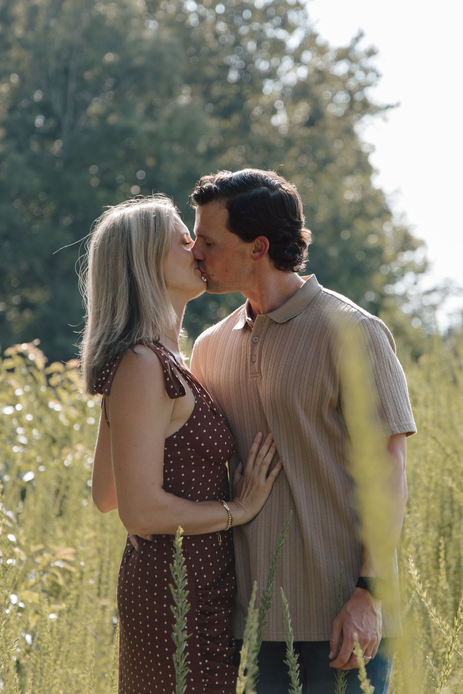A couple sharing a romantic kiss in a lush green field, woman in brown polka dot dress, man in striped polo shirt.