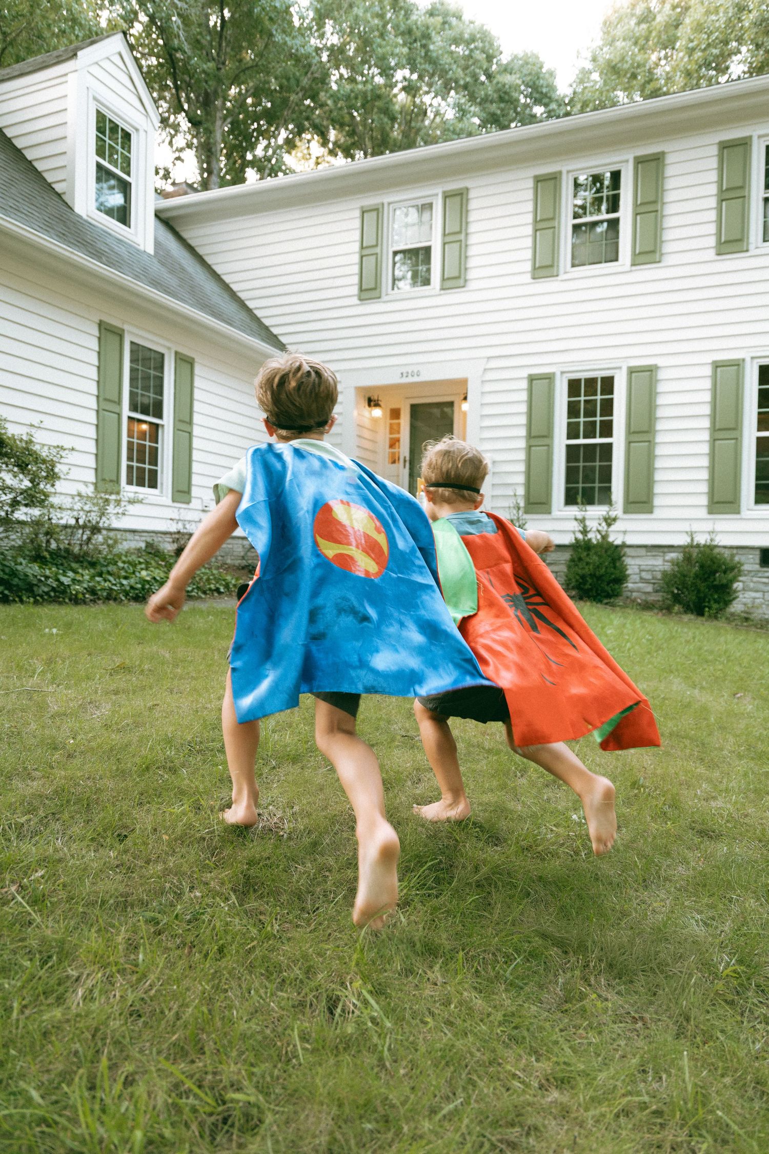 Two children wearing colorful superhero capes running barefoot across a green lawn toward a white colonial house.