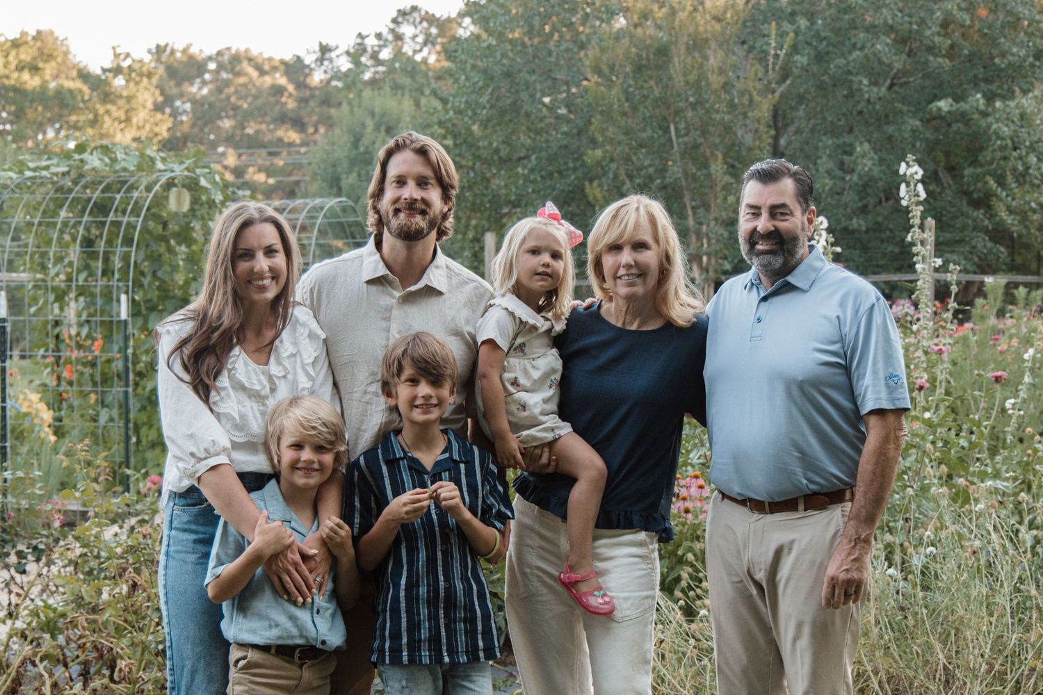 Multi-generational family of seven smiling together outdoors in a lush garden setting during a professional photo session.