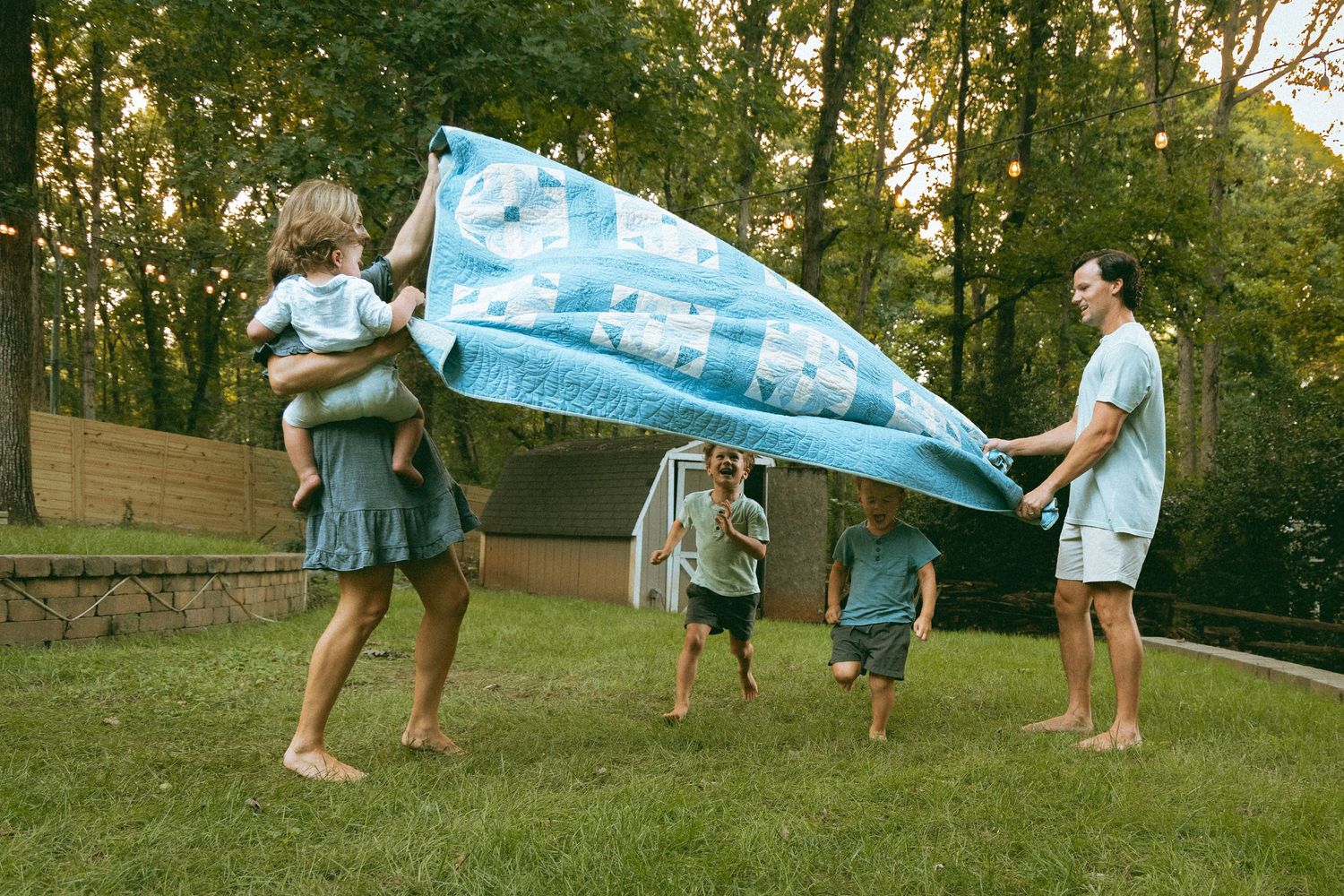 Family playing with a large blue blanket on green grass in a backyard at sunset, with children running underneath.