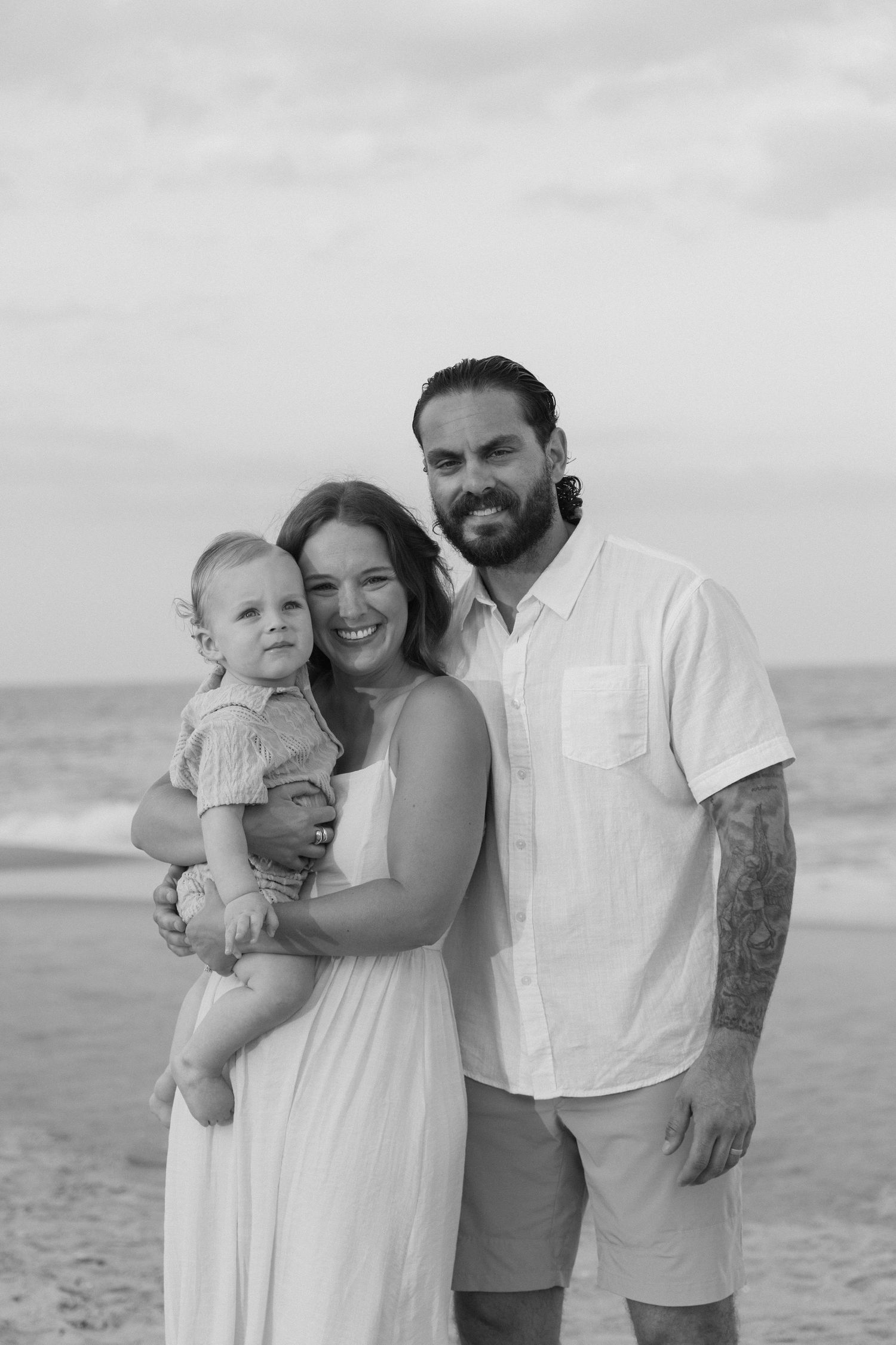 Black and white beach photo of smiling couple holding toddler, man with tattoos in white shirt, woman in white dress.