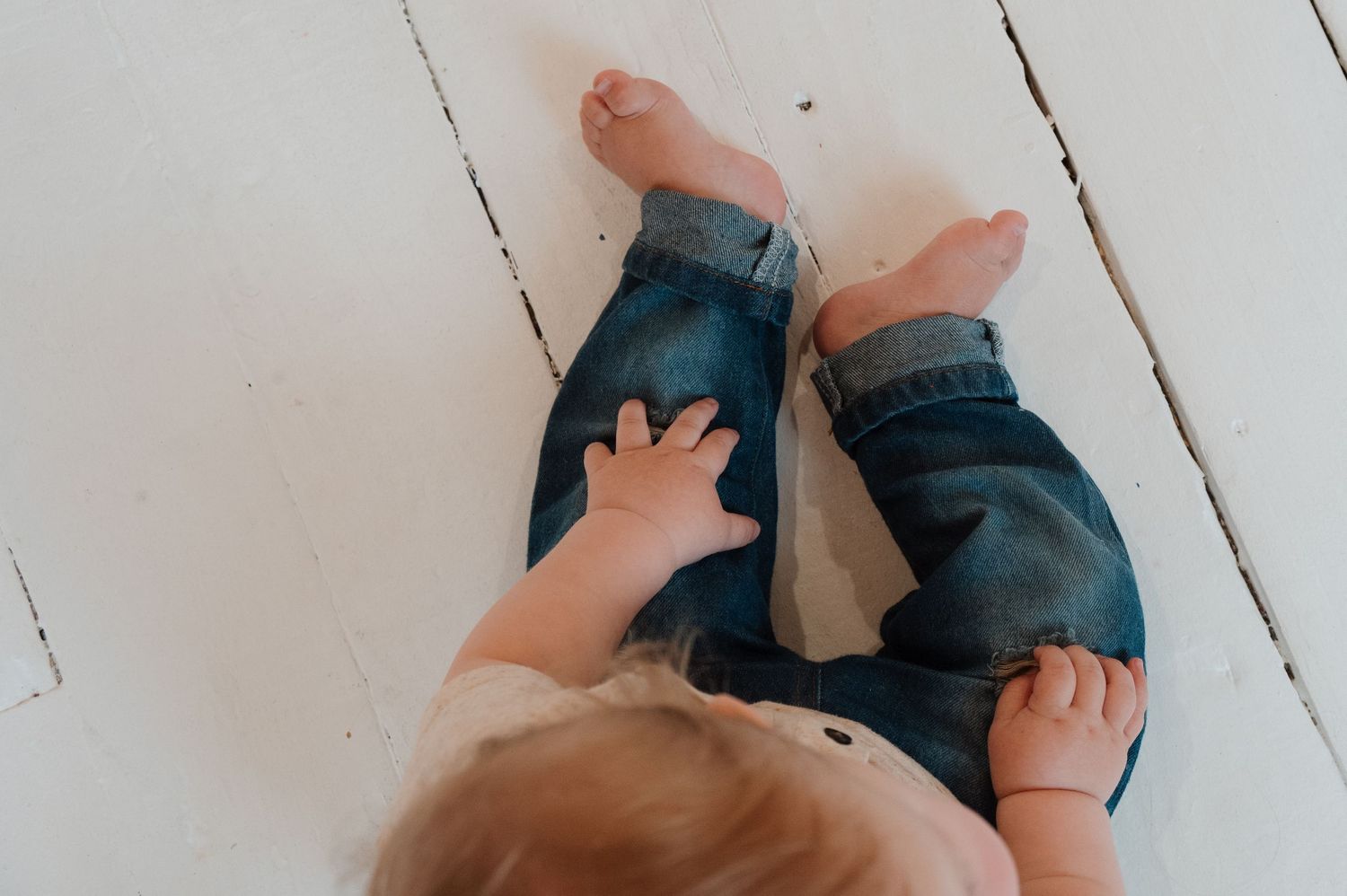 Overhead view of a baby sitting on white wooden floor, reaching toward adult legs in blue denim jeans.