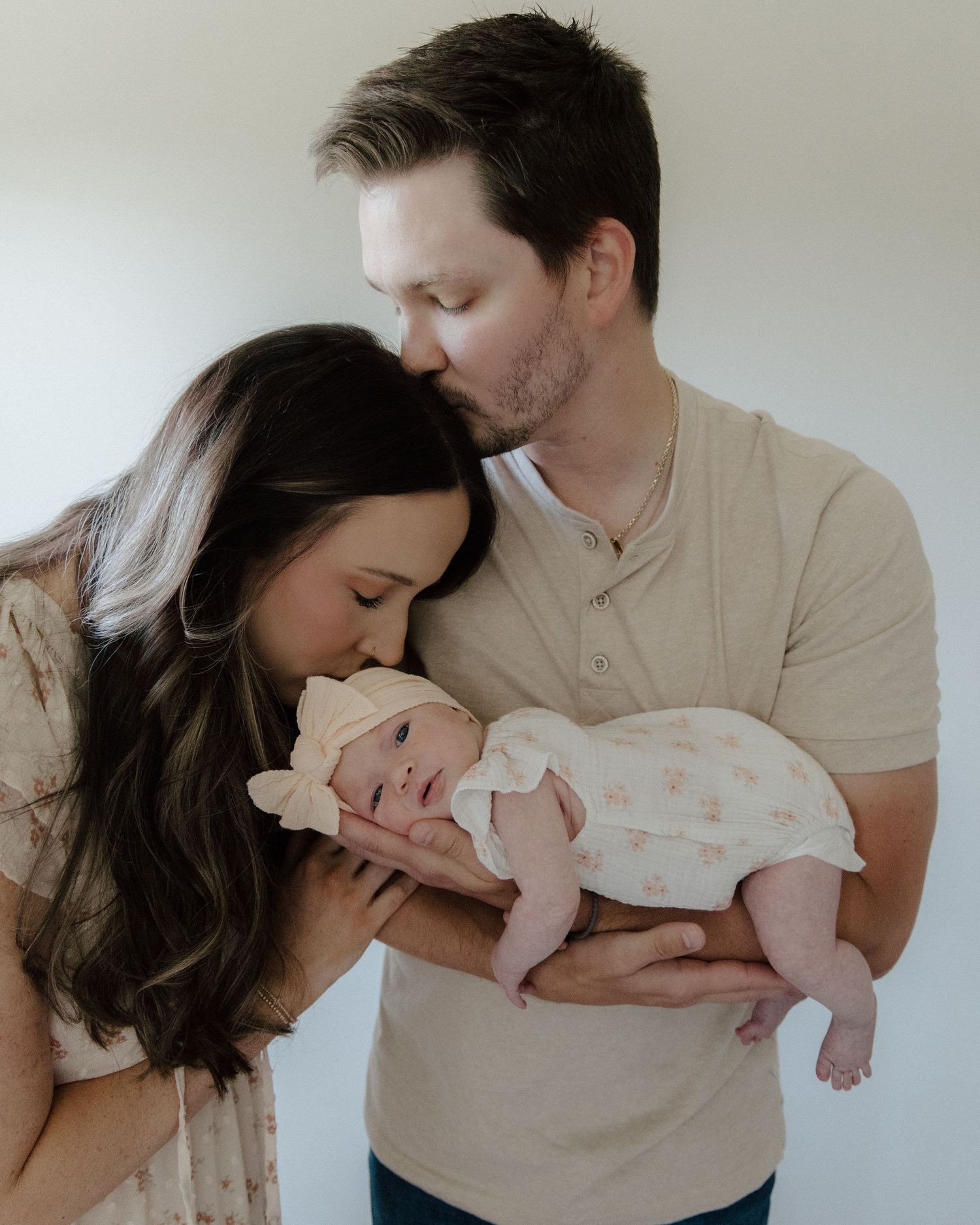A couple tenderly kisses their newborn baby, with the mother leaning in and father holding the infant in a soft studio setting.