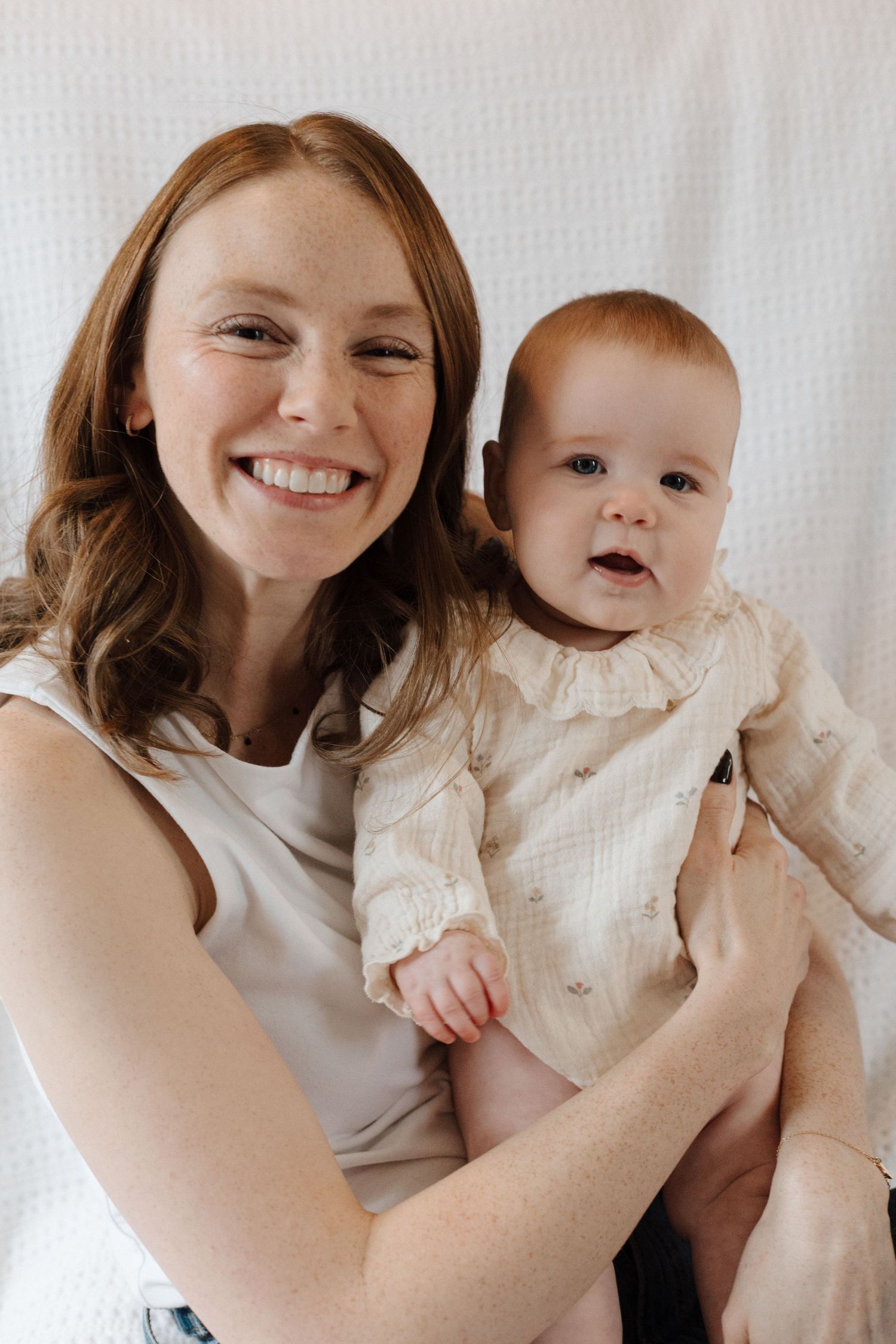 Smiling redhead woman holding an adorable baby in a cream ruffle outfit against a white background.
