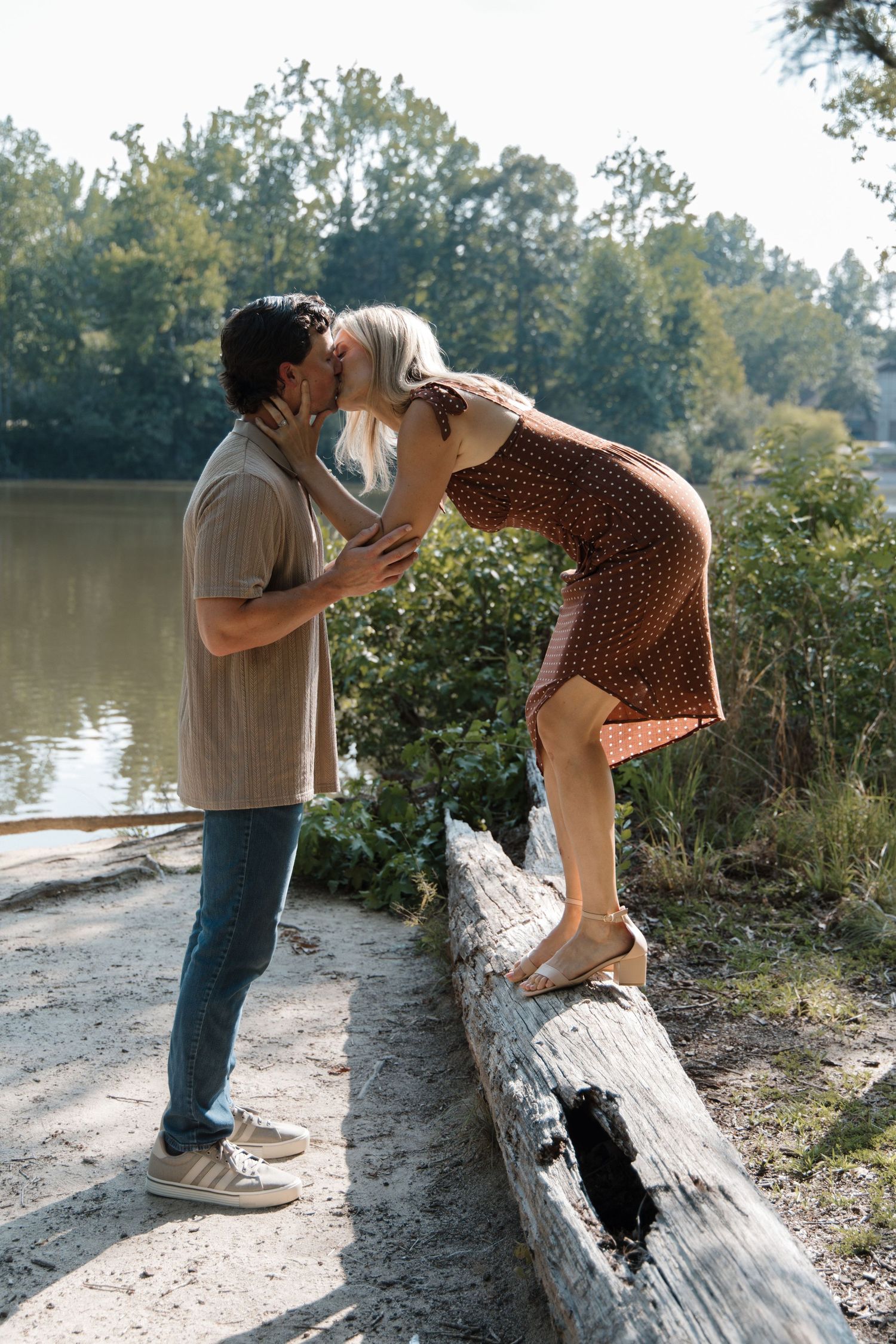 Couple sharing a romantic kiss outdoors, woman standing on a log by a river, surrounded by lush green trees.