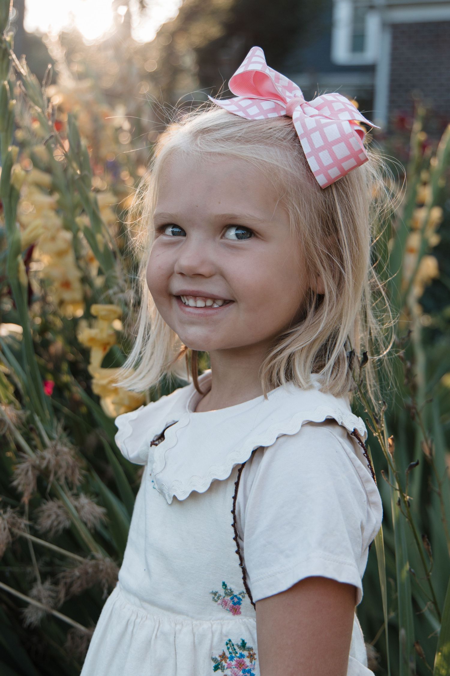 Smiling blonde girl with pink bow hair clip wearing white floral dress standing among yellow flowers in garden.