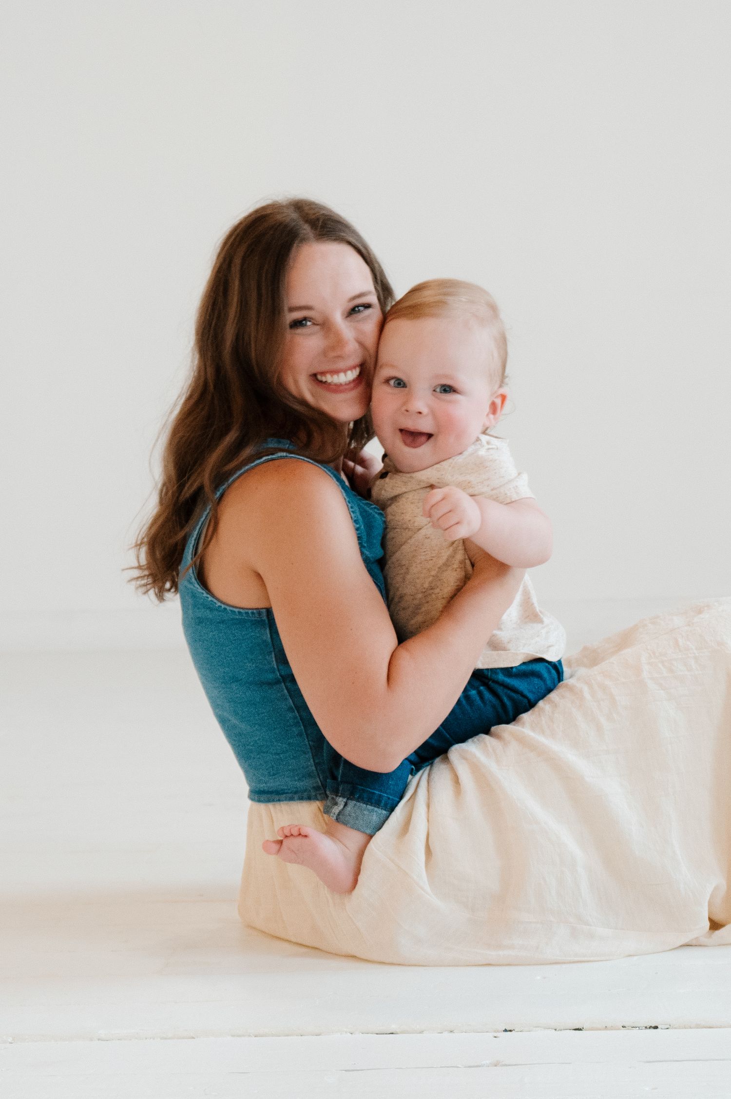 Smiling mother sitting on white floor hugging happy baby in bright studio portrait photo.