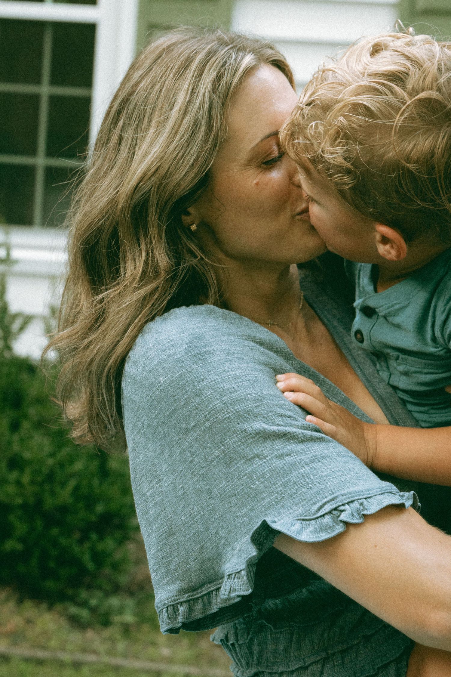 A mother tenderly kisses her young blond curly-haired child outdoors near a house.