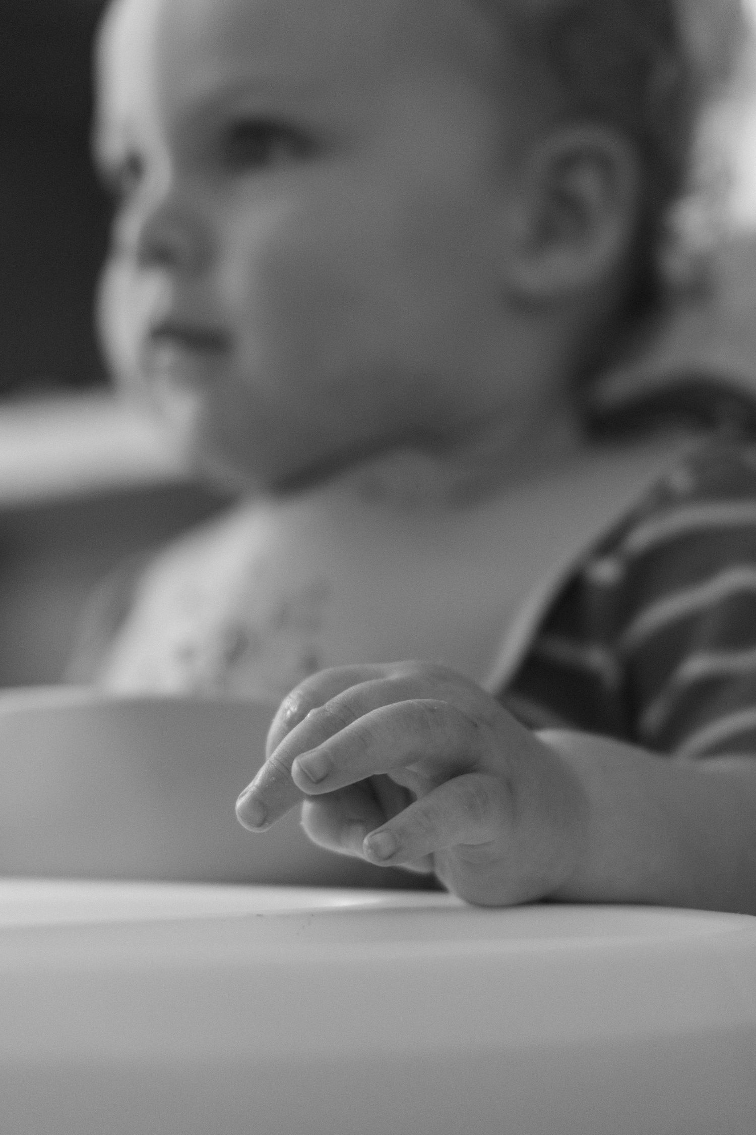Black and white photo of a baby resting tiny hand on white table, with soft focus face in background.