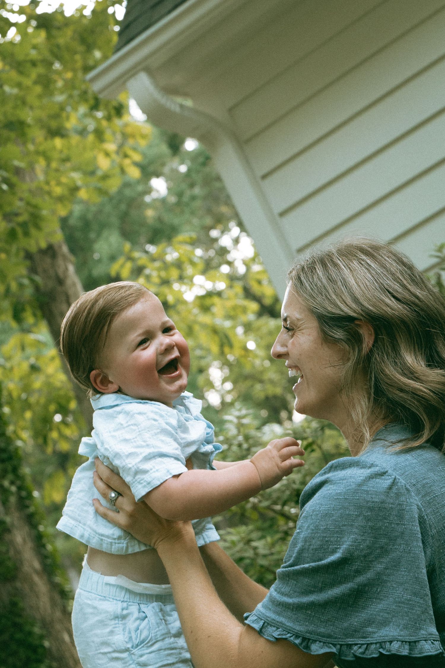 A joyful mother holds her laughing baby outdoors near a white house surrounded by green trees.
