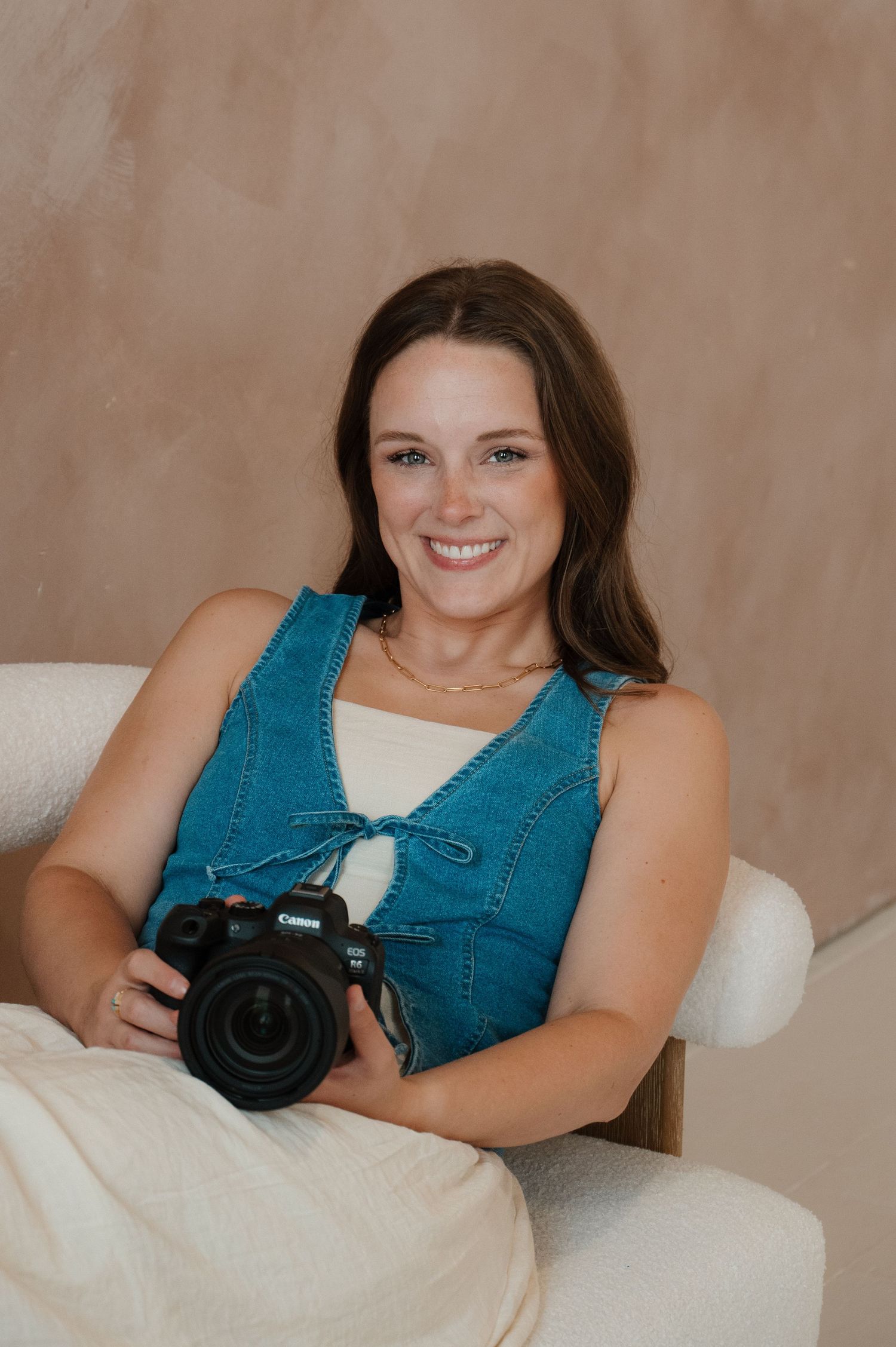 Smiling young woman holding a Canon DSLR camera, wearing a denim vest, seated in a white chair against a beige backdrop.