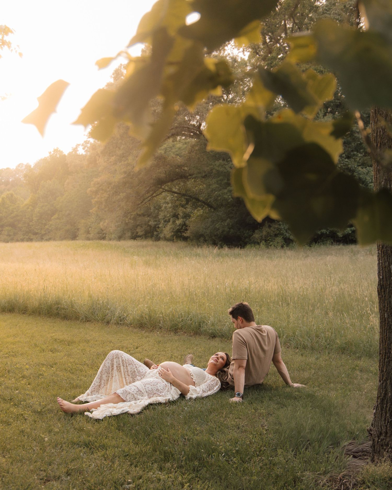 A couple relaxes in a sunlit meadow, woman in white lace dress lying down, man seated beside her under a tree.