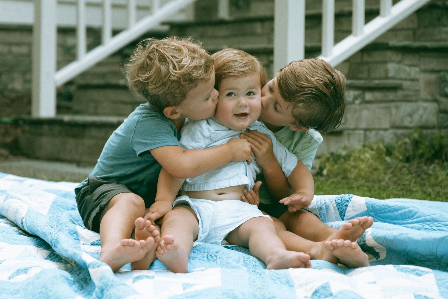 Three toddlers sitting on a blanket outdoors, two children kissing a smiling baby on the cheeks.