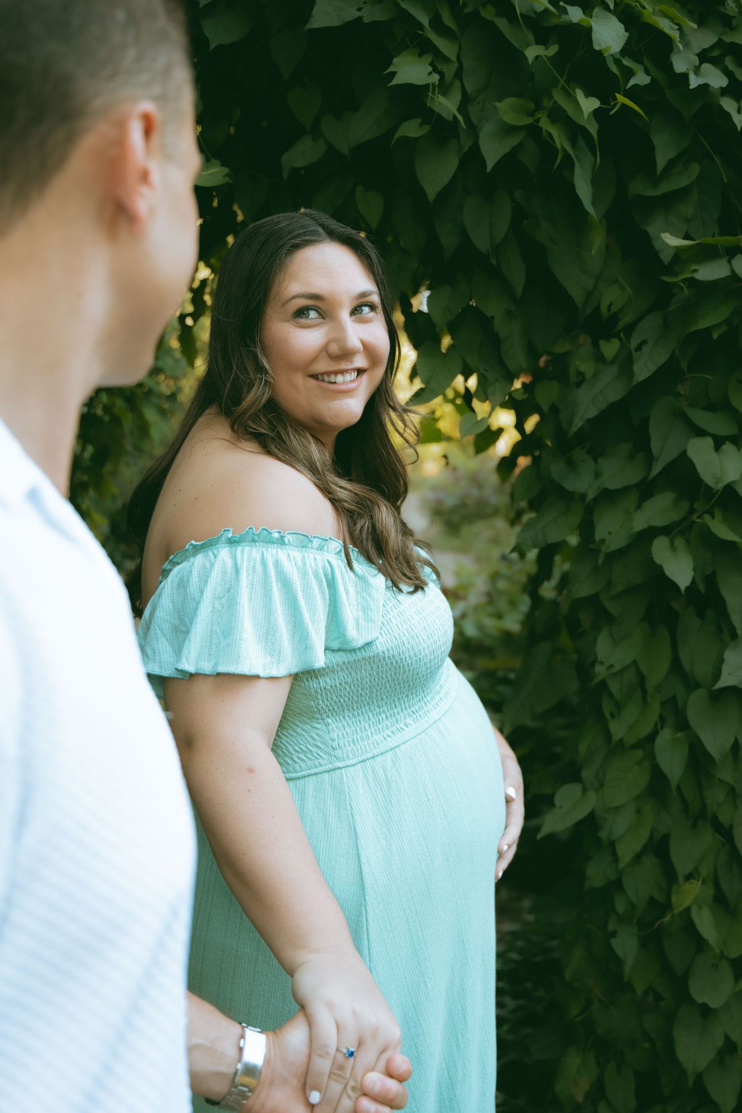 Pregnant woman in mint off-shoulder dress holds hands with partner outdoors among lush green foliage.