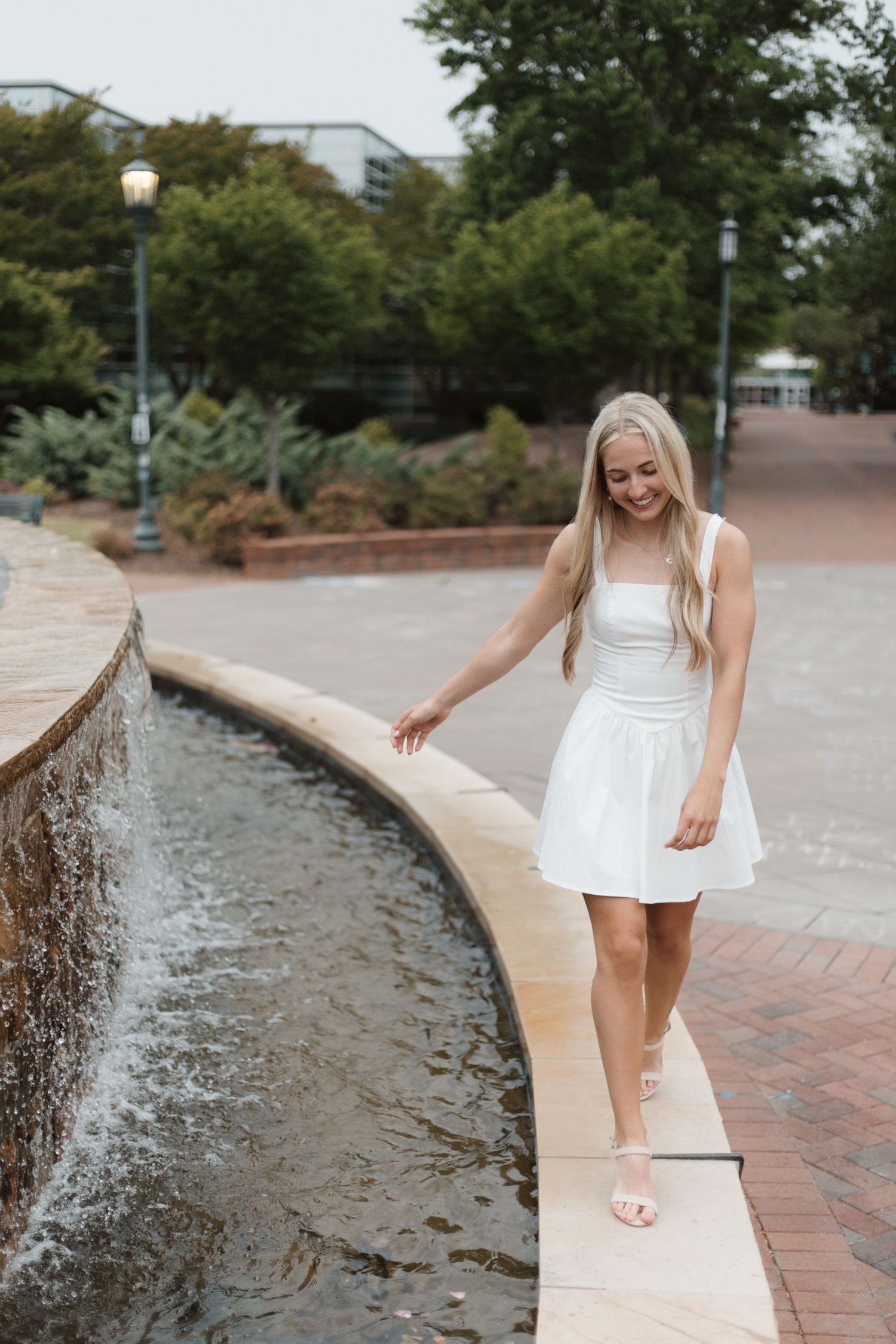 Young blonde woman in white dress walking along fountain edge in urban park setting.