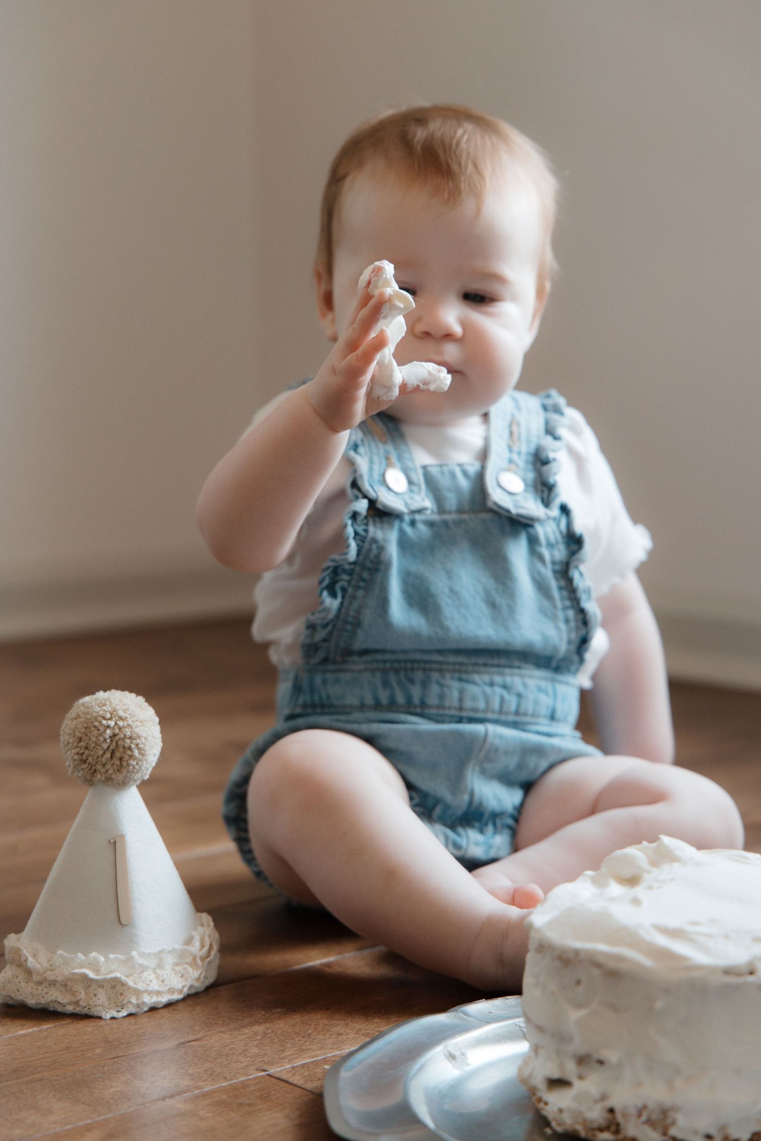 Baby in denim overalls smashing first birthday cake on wooden floor with party hat nearby.