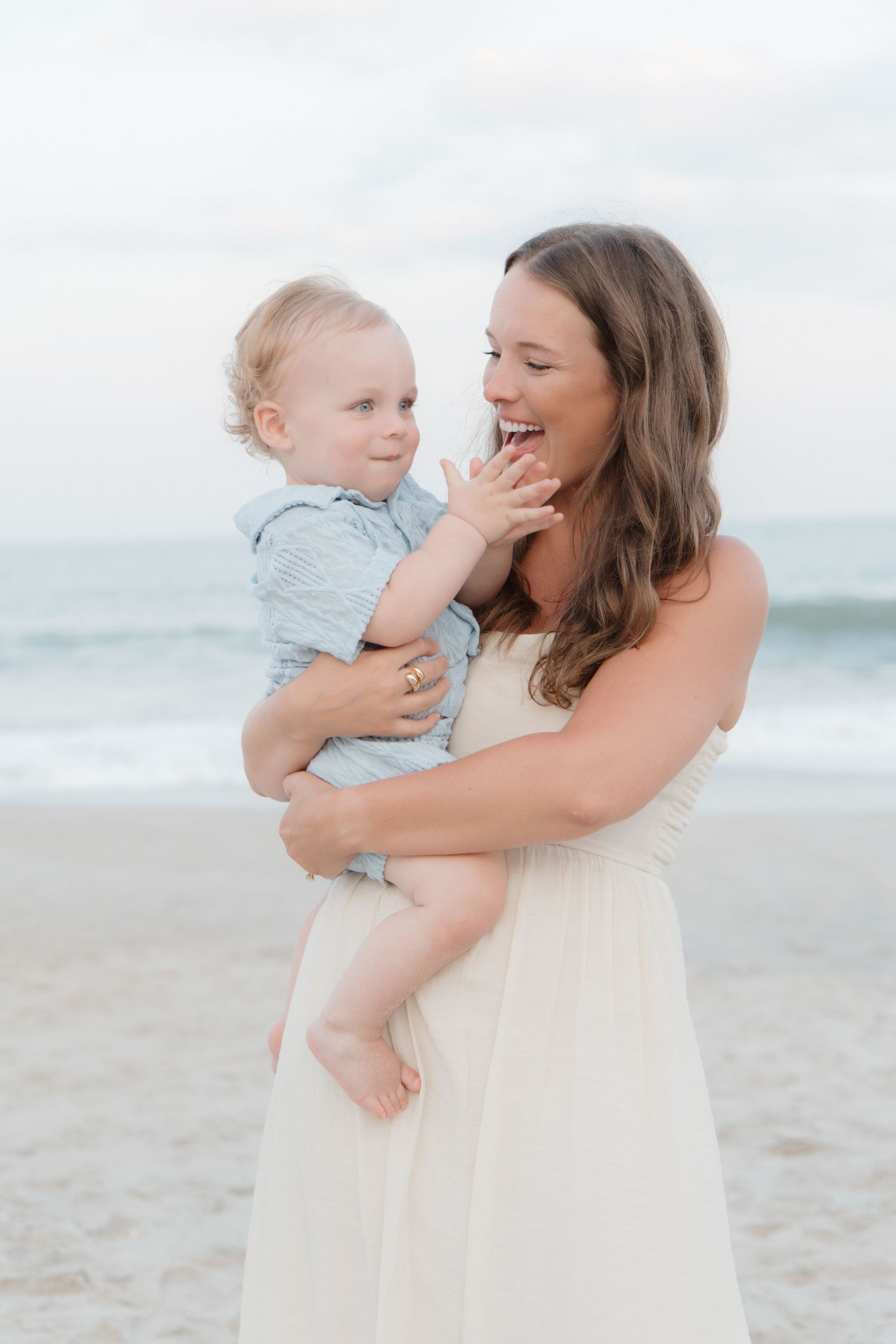 A smiling mother in a white dress holds her baby boy on a beach, sharing a joyful moment by the ocean.
