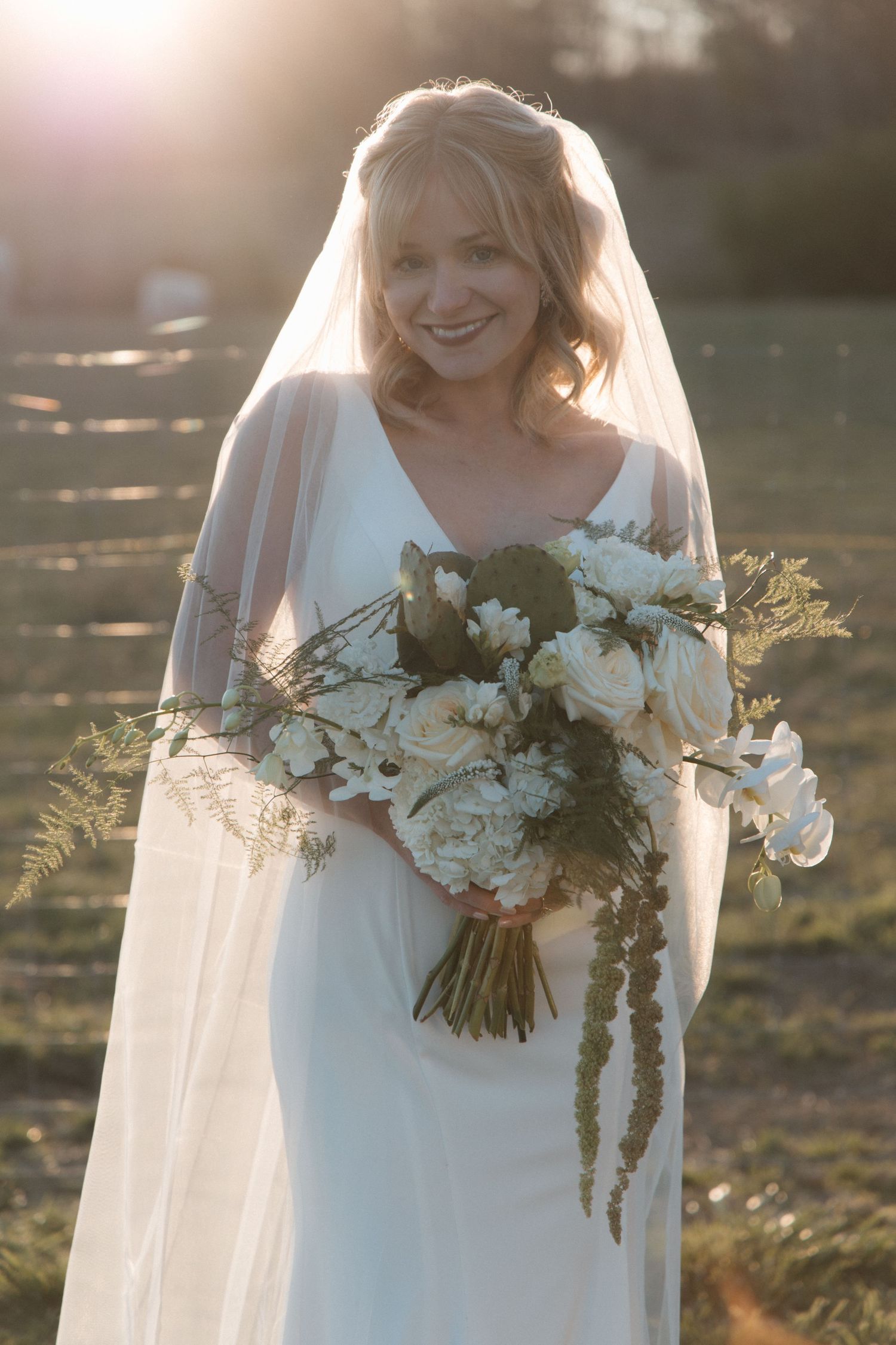 Smiling bride in white gown and veil holds white floral bouquet with greenery, bathed in golden sunset light outdoors. CHARLOTTE NC