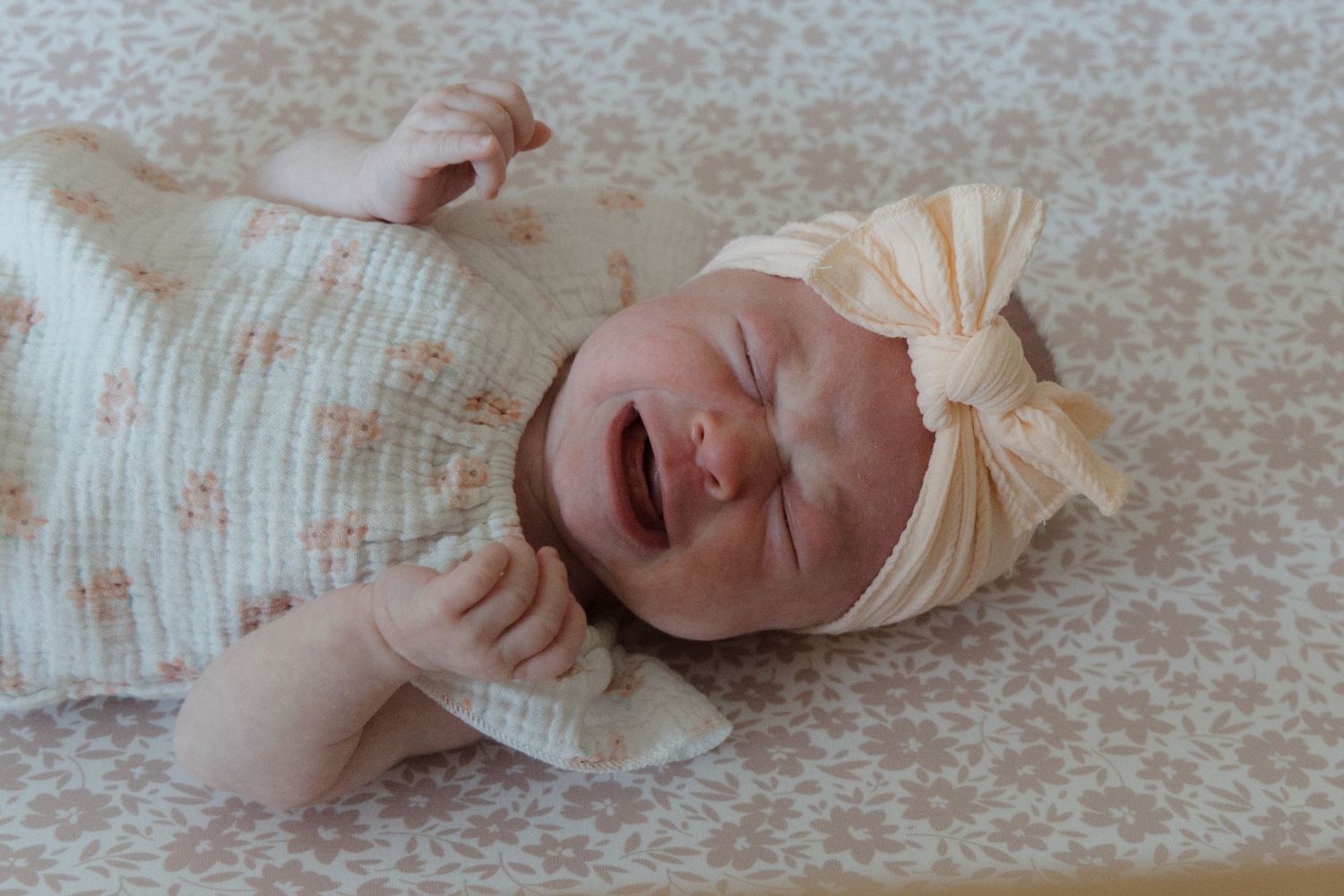Newborn baby girl crying in floral outfit with cream bow headband, lying on soft patterned blanket.