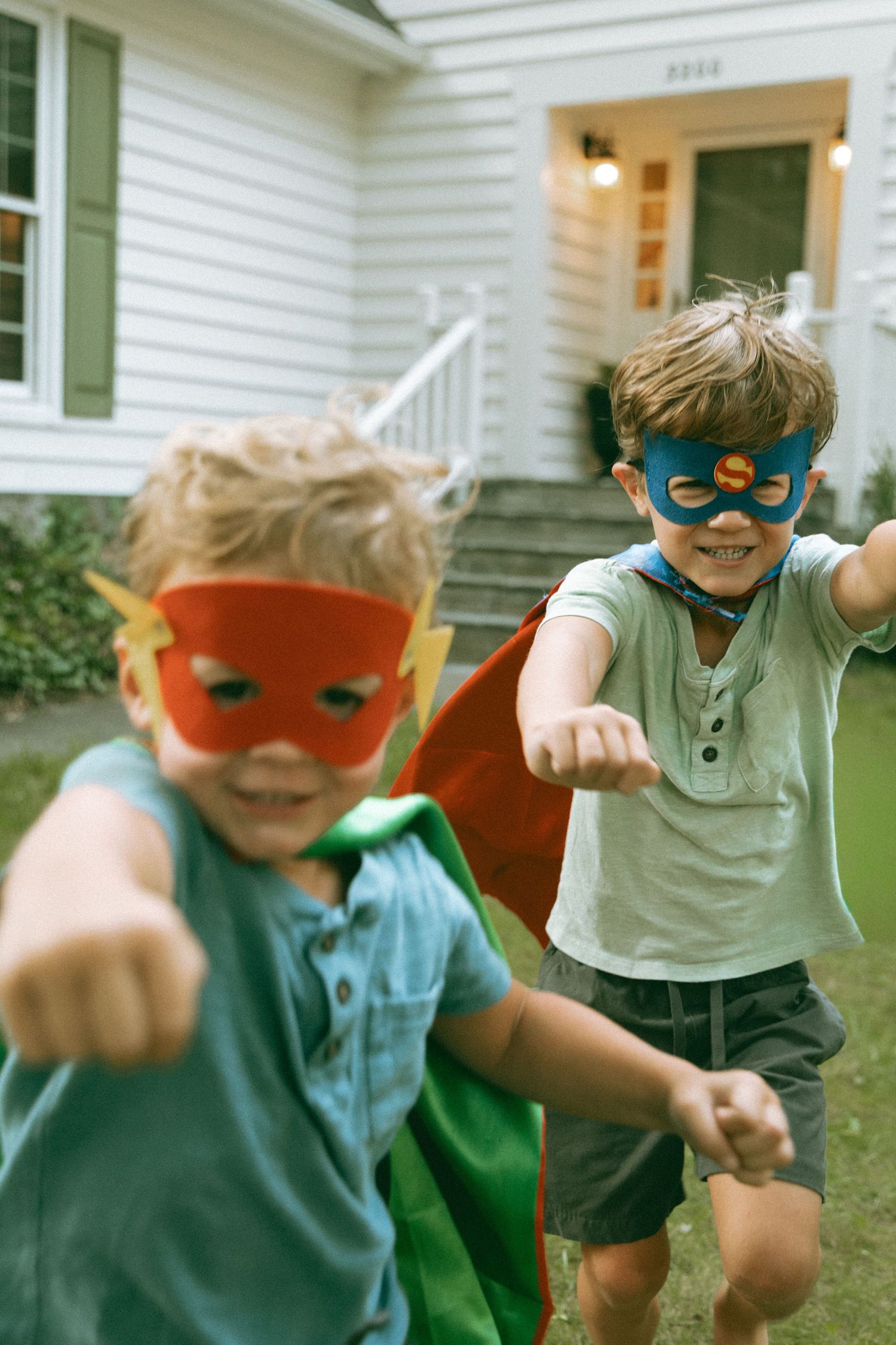 Two young boys wearing superhero masks and cape running and playing outside home.