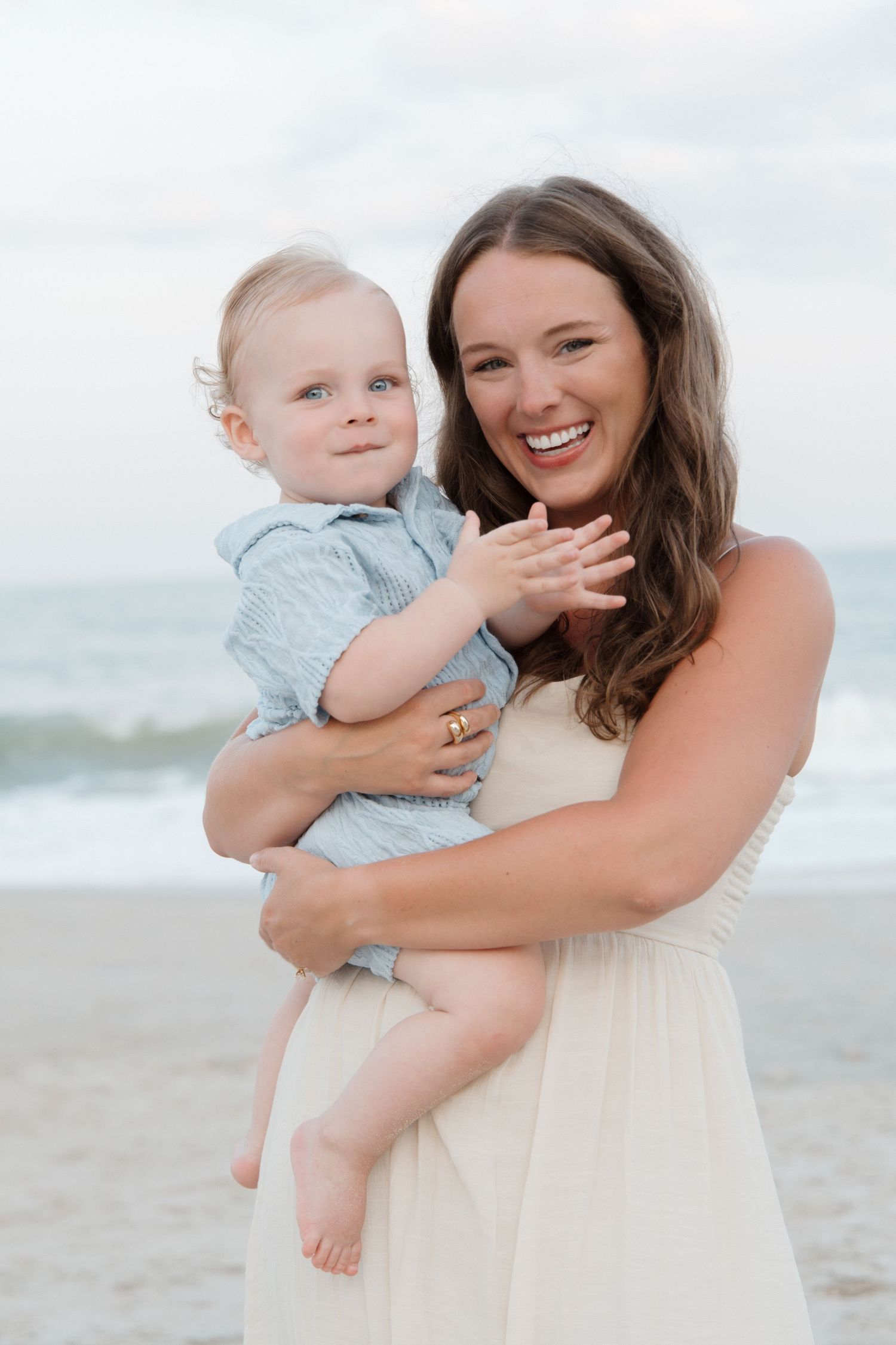 Smiling mother holding her baby boy on a beach with ocean waves in the background.