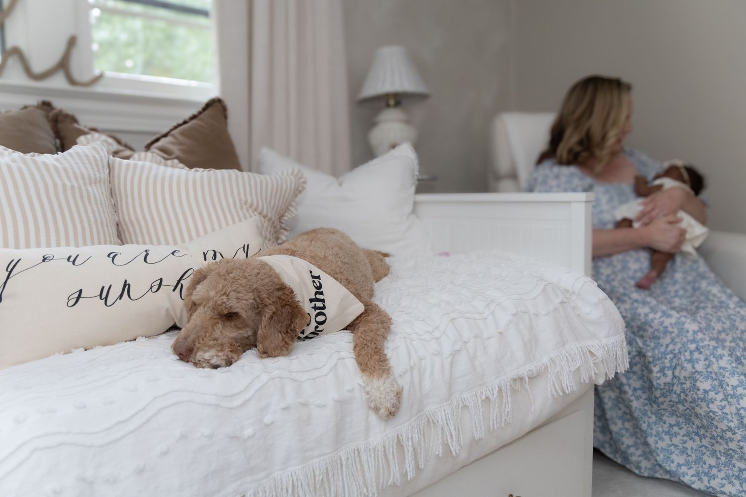 A golden doodle dog rests on a white bed while a woman in a floral dress holds a newborn baby in a cozy bedroom.