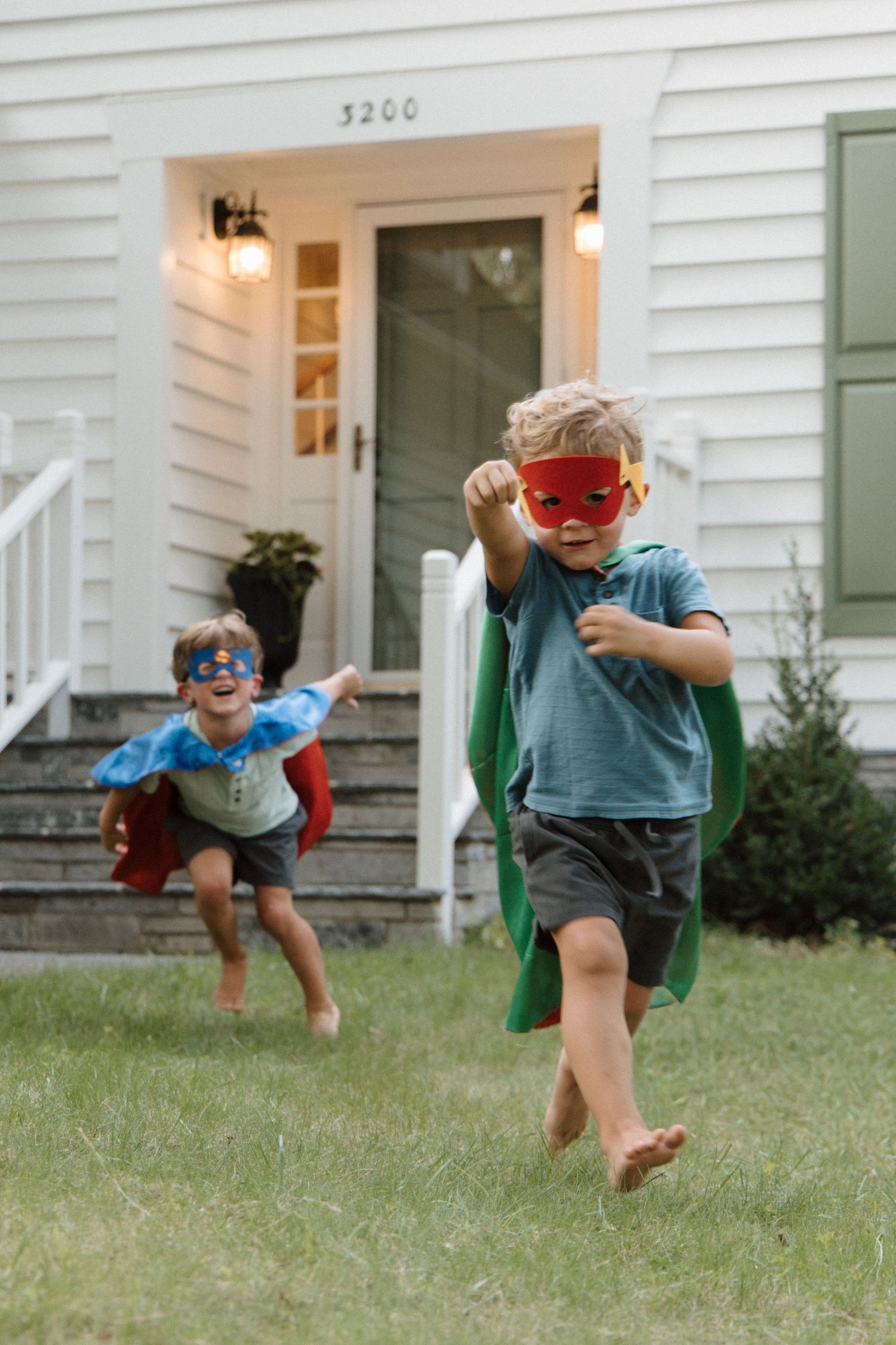 Two children in superhero capes and masks running barefoot on grass in front of a white house.