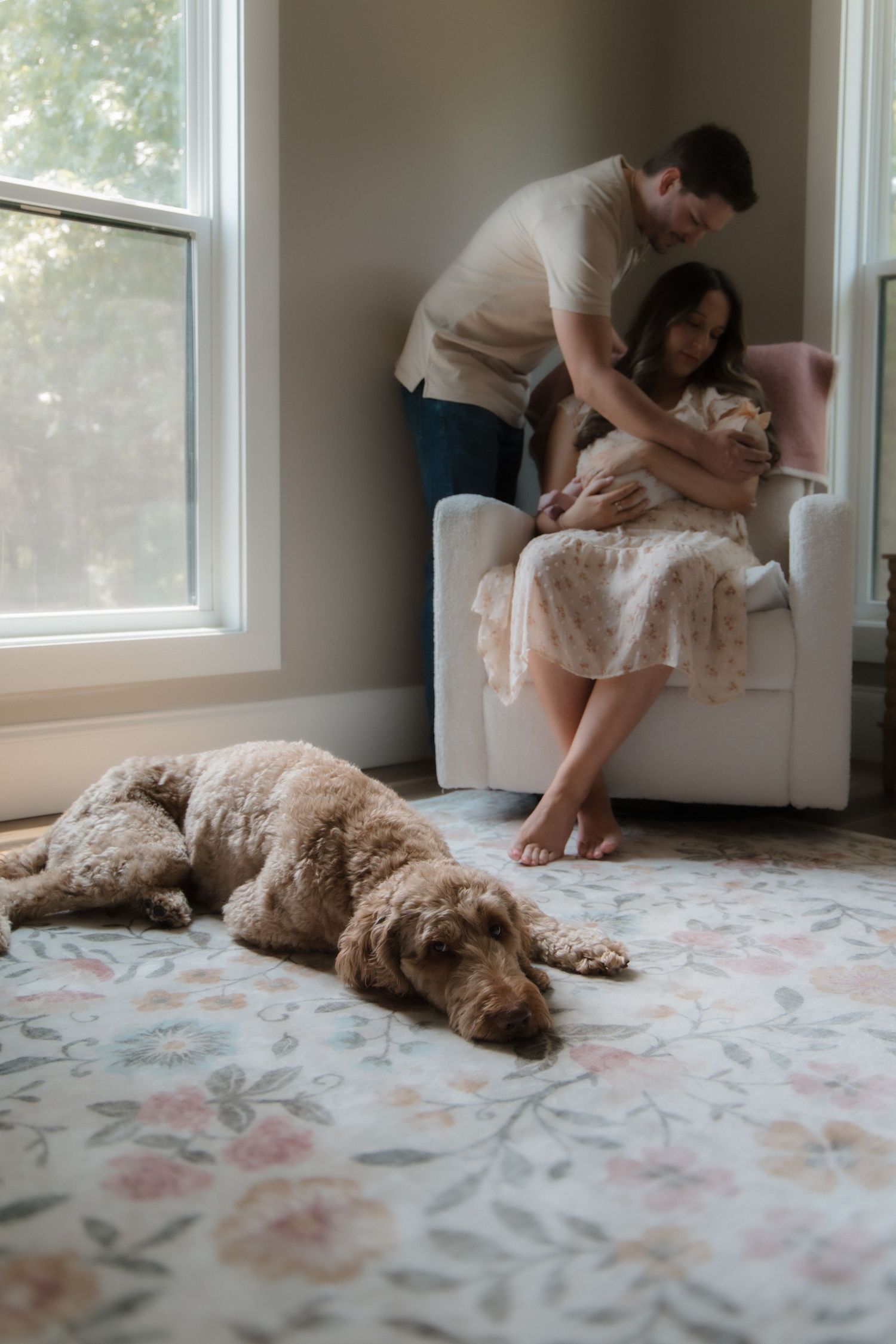 A couple with a newborn baby in a chair while their golden doodle dog rests on a floral rug nearby.
