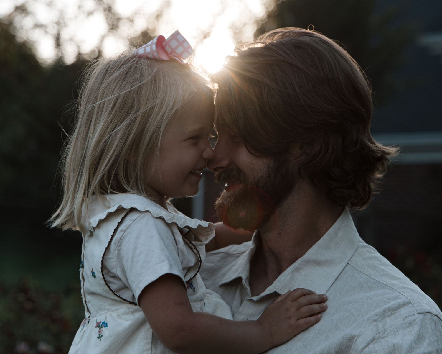 A father holds his young blonde daughter with a bow, touching foreheads and smiling in warm sunset backlight.