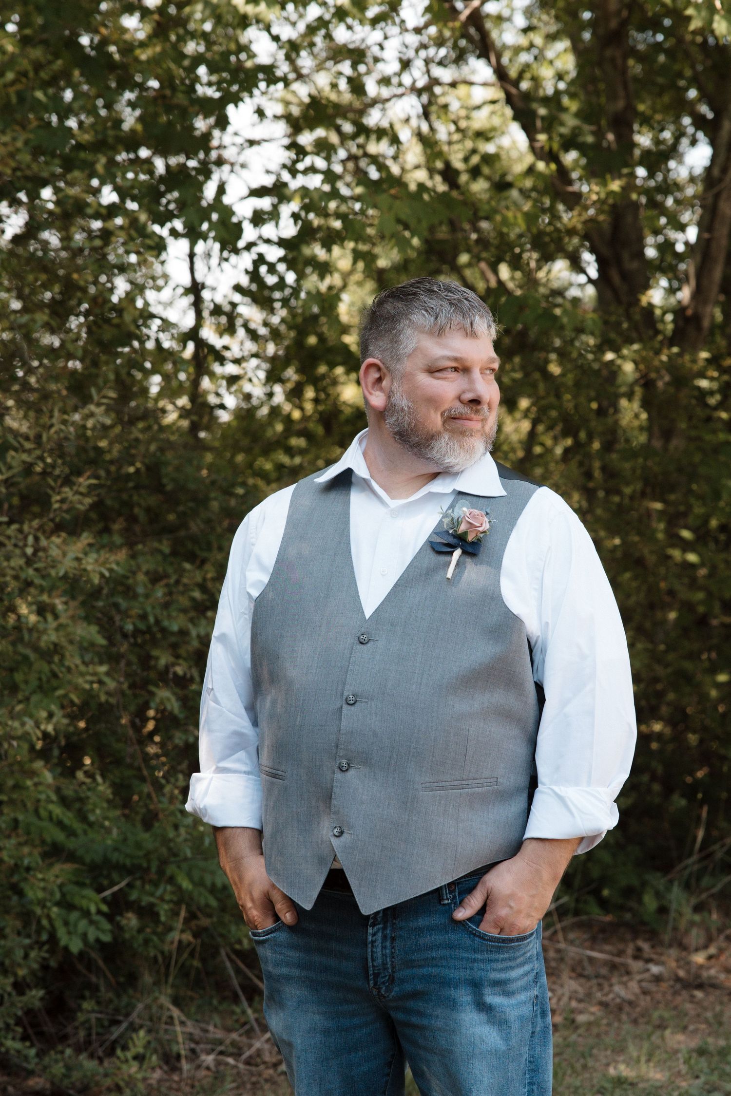 Middle-aged groom in gray vest, white shirt, and jeans with boutonniere poses outdoors among green trees.