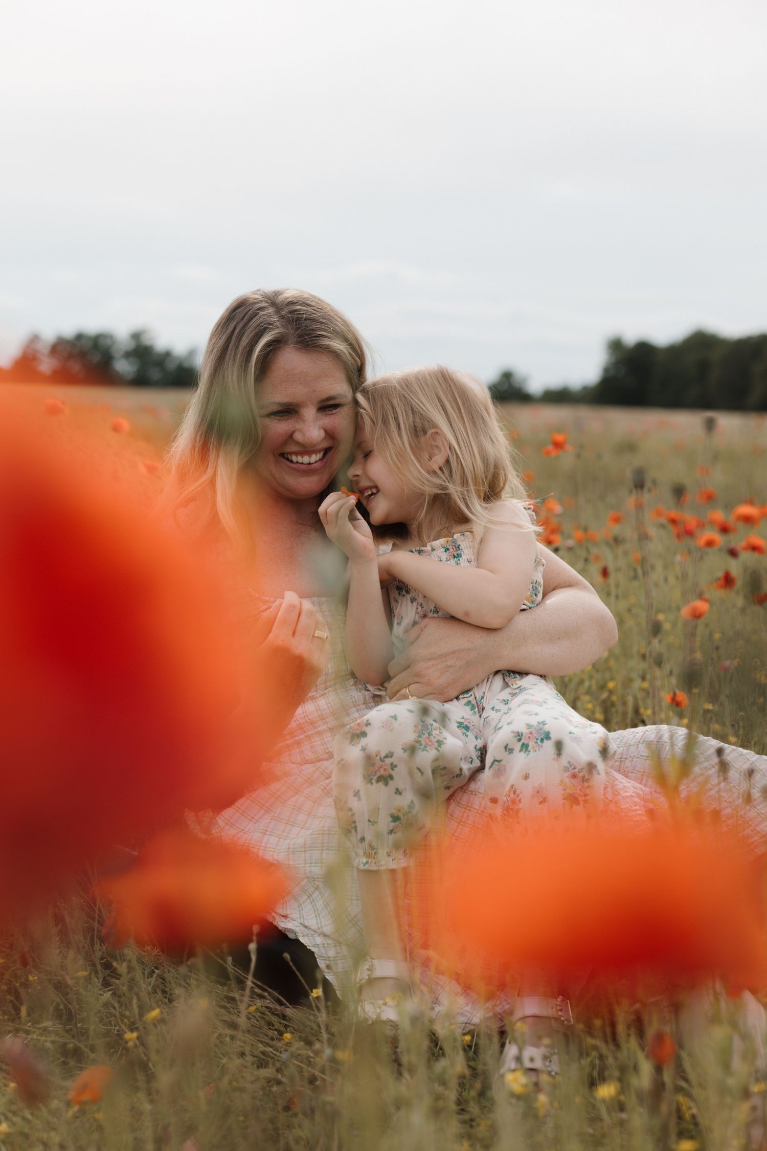 A mother and young daughter laugh together while sitting in a vibrant red poppy field on an overcast day. CHARLOTTE NC PHOTOGRAPHER