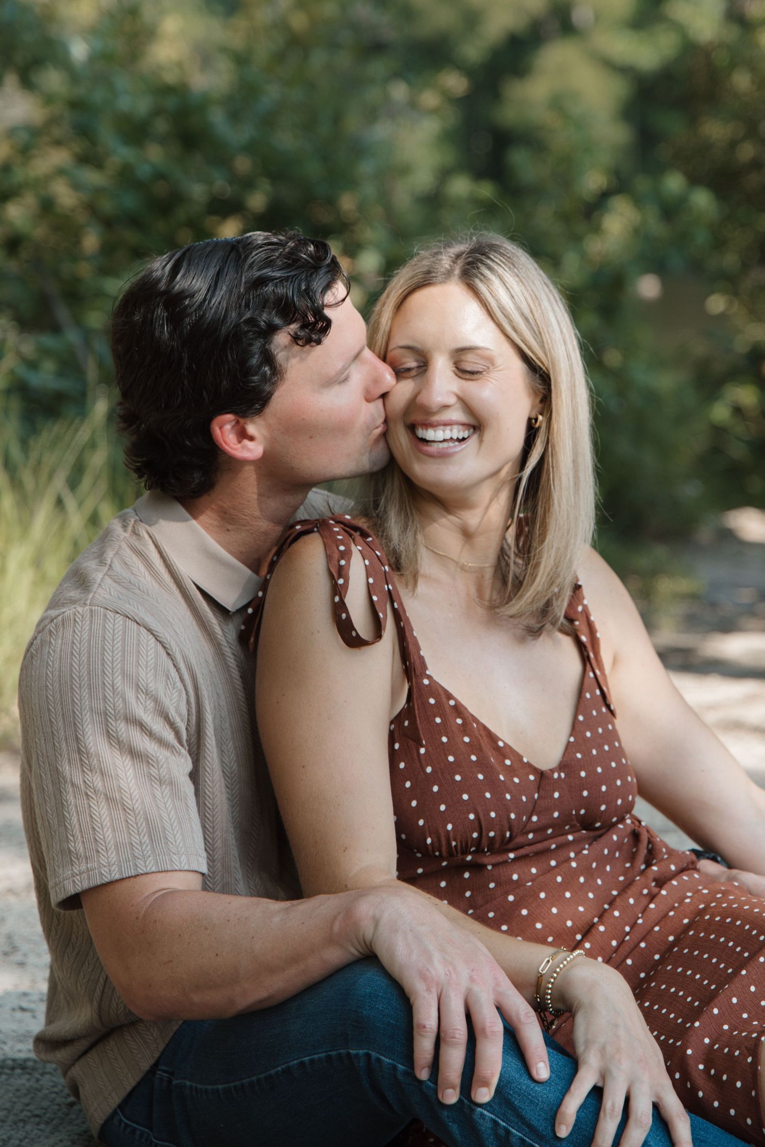 A man kisses a laughing blonde woman on the cheek outdoors during a romantic couples photo session.