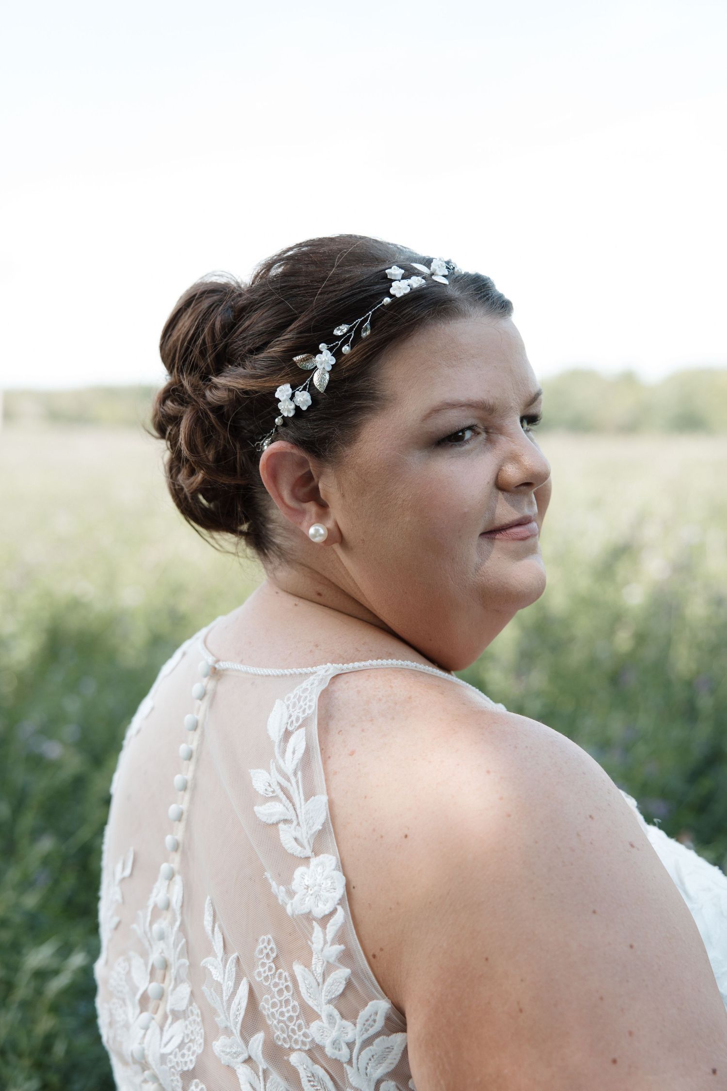 Bride in white lace gown and floral hair vine looks over shoulder in outdoor field setting.