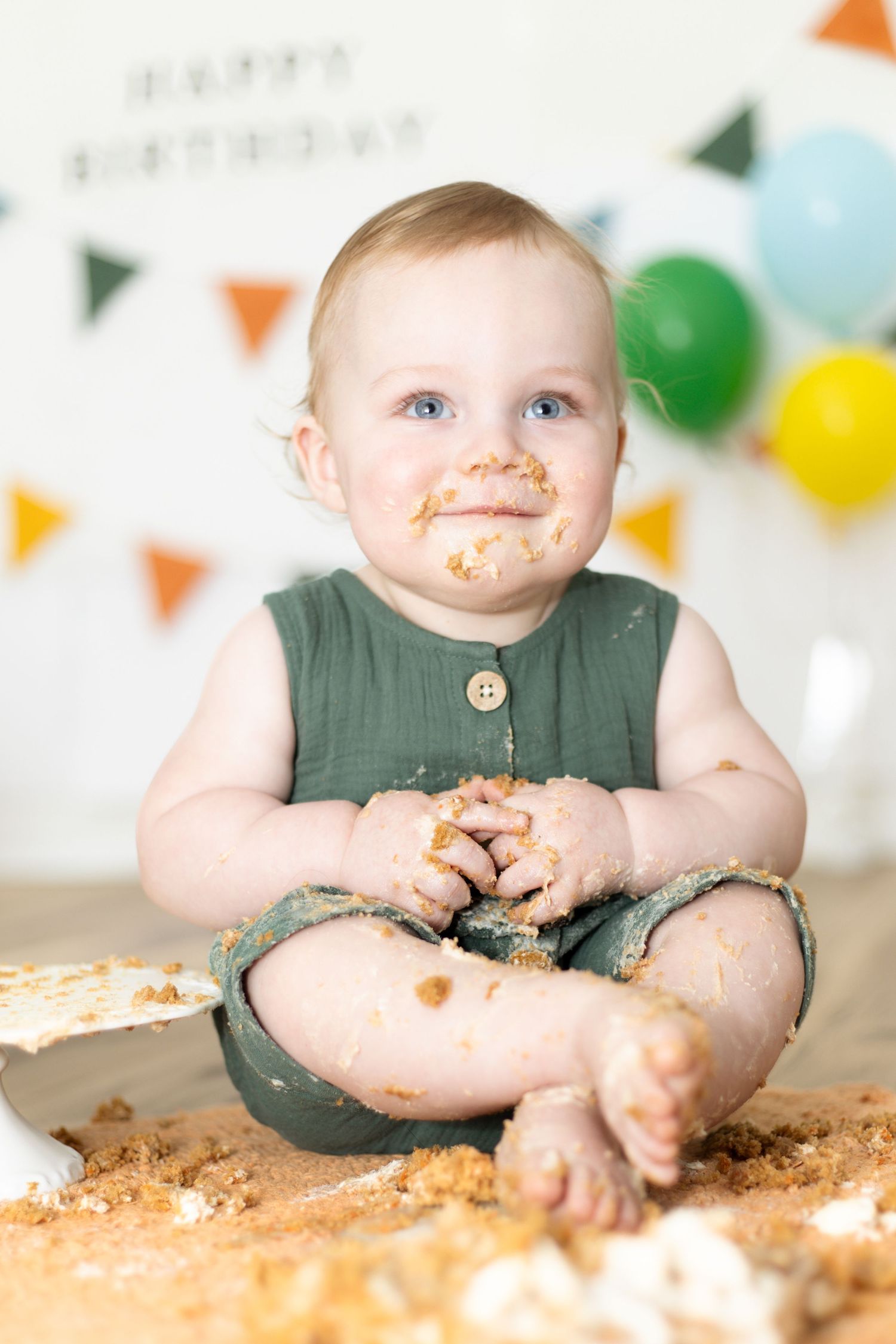 A messy baby with cake-covered face and hands sits on the floor at a first birthday cake smash celebration.