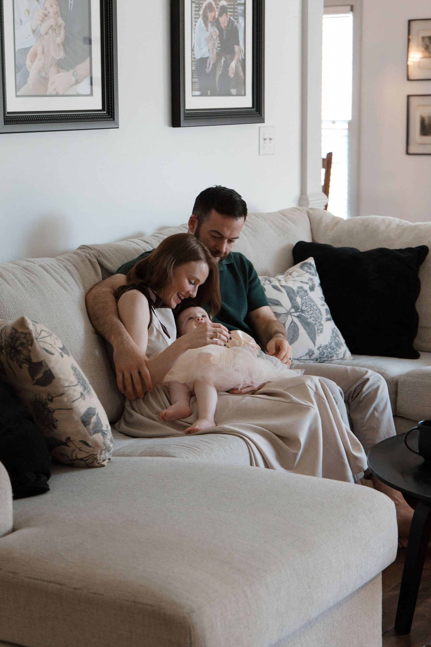 Young couple sitting on a beige sofa holding their newborn baby in a cozy living room with framed photos.
