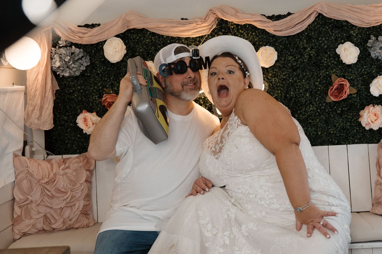 Bride and groom in white outfits celebrating at wedding reception photo booth with floral wall backdrop.
