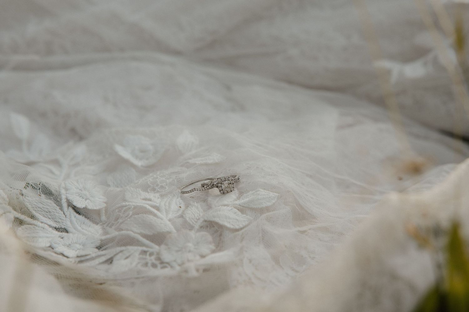Diamond engagement ring resting on delicate white lace wedding dress fabric with floral embroidery details.