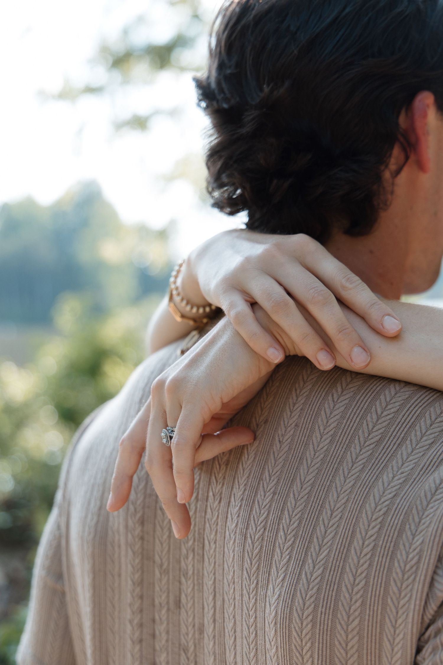 A woman's hand with an engagement ring and bracelet resting on a man's shoulder in a tender embrace.