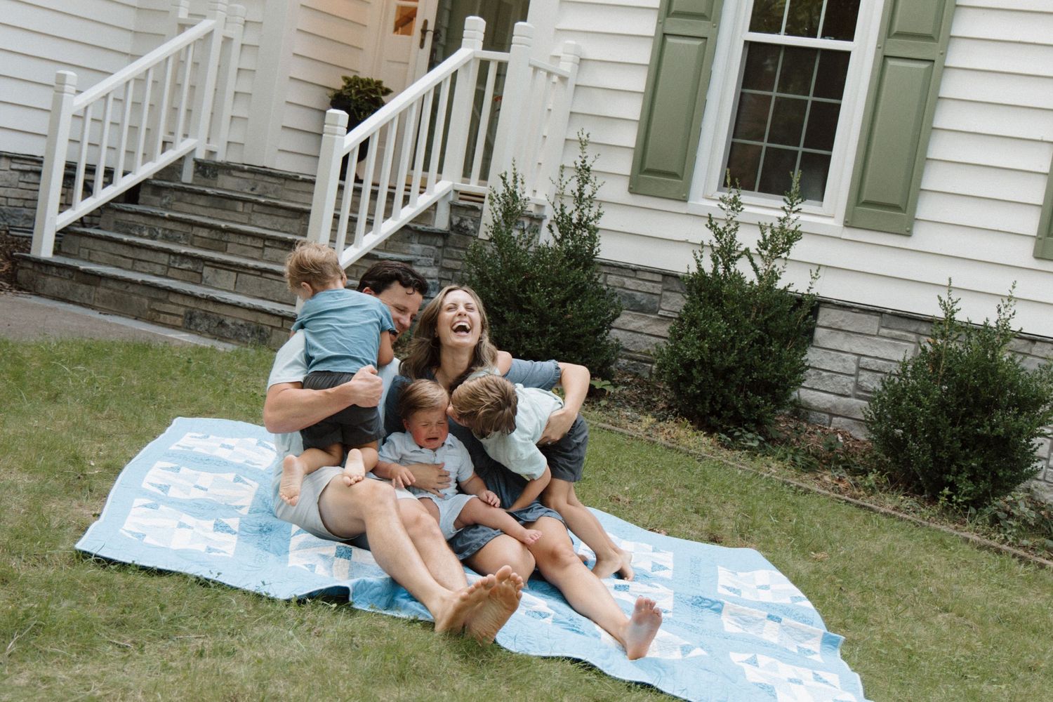 Happy family laughing and playing together on a blue blanket on the front lawn of a white house.