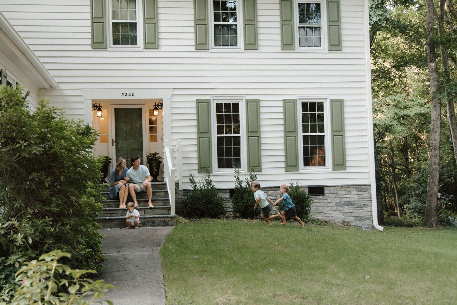 Family relaxing on front porch steps of white colonial home with green shutters and lush lawn.