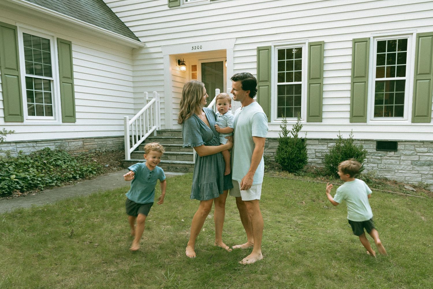 Happy family with three young children playing in front yard of white house with green shutters on sunny day.